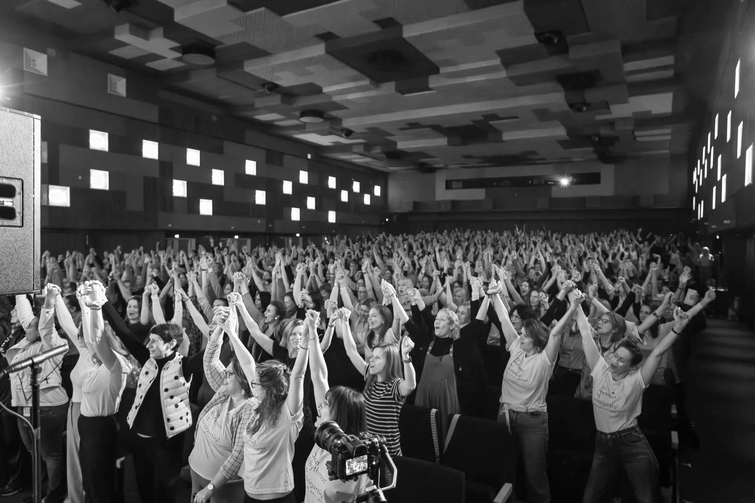 Menschen in einem Konzertsaal heben zusammen die Hände und scheinen zu feiern.