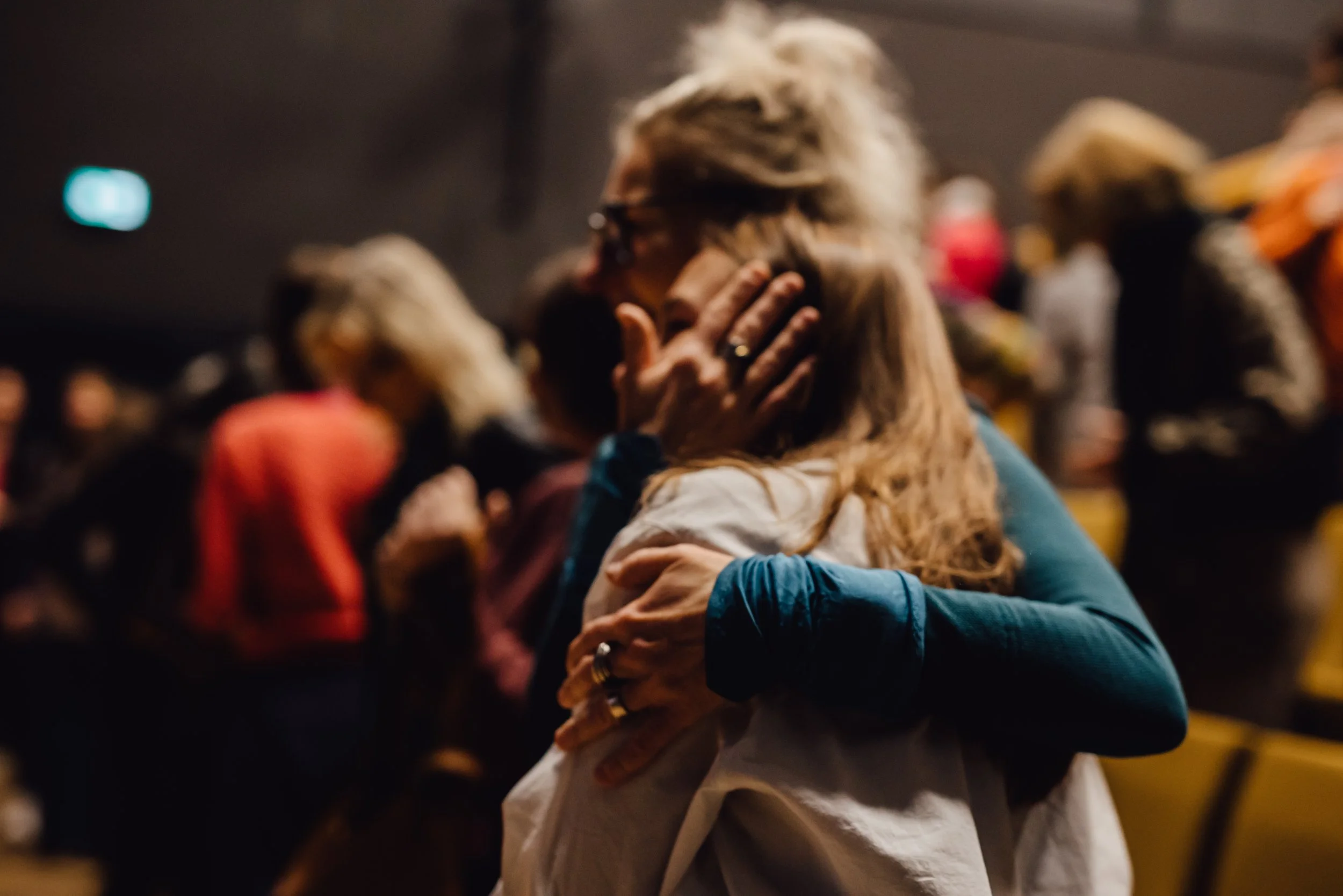 Zwei Frauen umarmen sich in einer Menschenmenge, halten sich an den Schultern und Rücken, in einem dunklen Raum mit gelben Sitzplätzen.