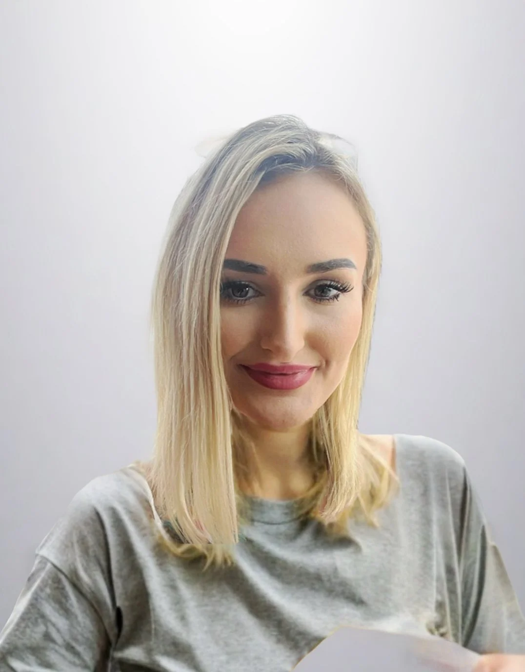 A woman with shoulder-length blonde hair, makeup, and a gray shirt, smiling against a plain white background.