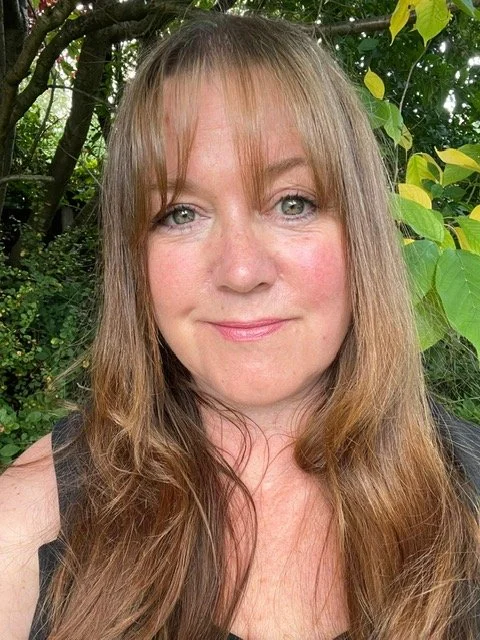 A woman with long blonde hair and blue eyes smiling outdoors with green foliage in the background.