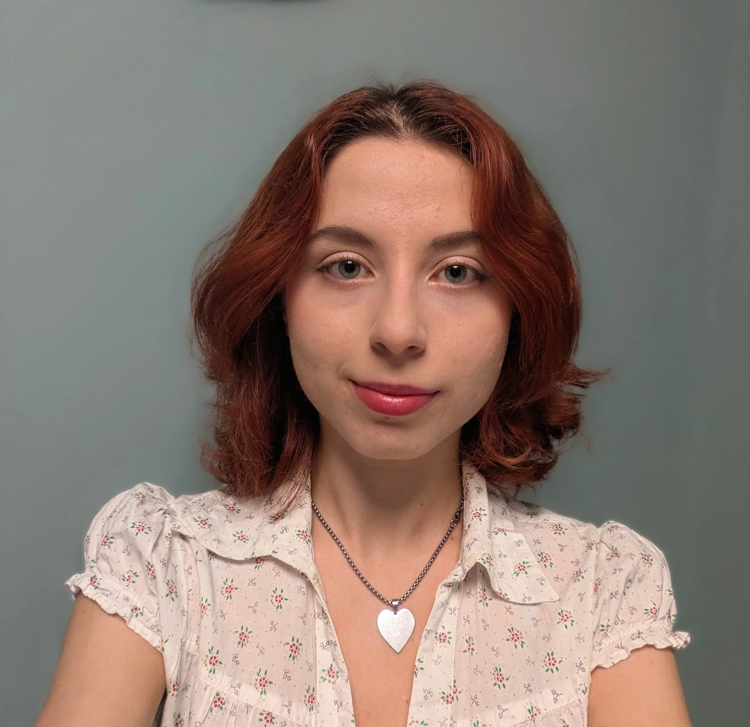 A woman with shoulder-length red hair, light skin, wearing a white blouse with a floral pattern and a silver heart-shaped pendant necklace, smiling softly against a muted gray background.