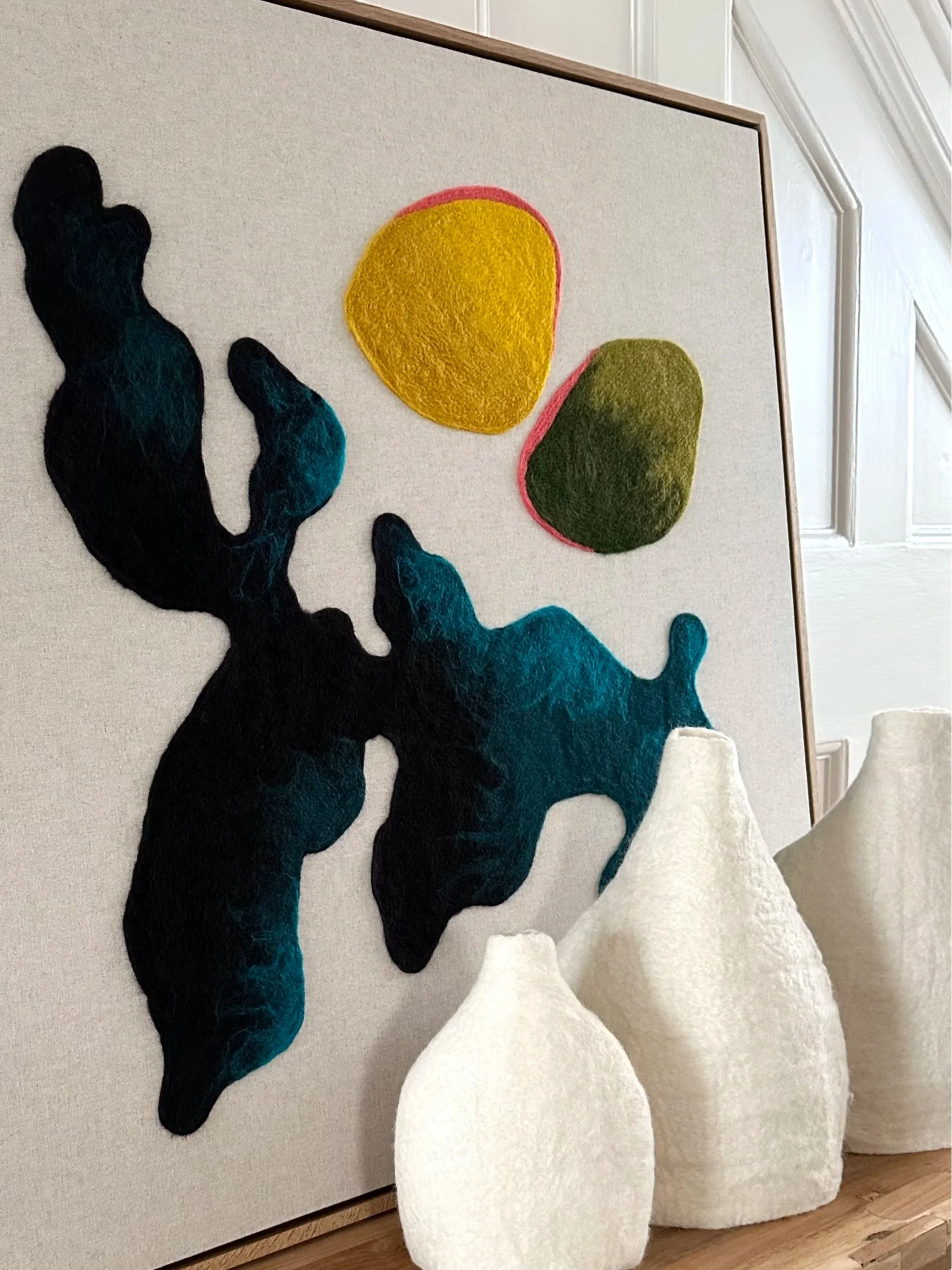 An abstract fabric art piece featuring organic shapes in dark blue, teal, yellow, and green, along with three standing white textured vases placed in front of it.