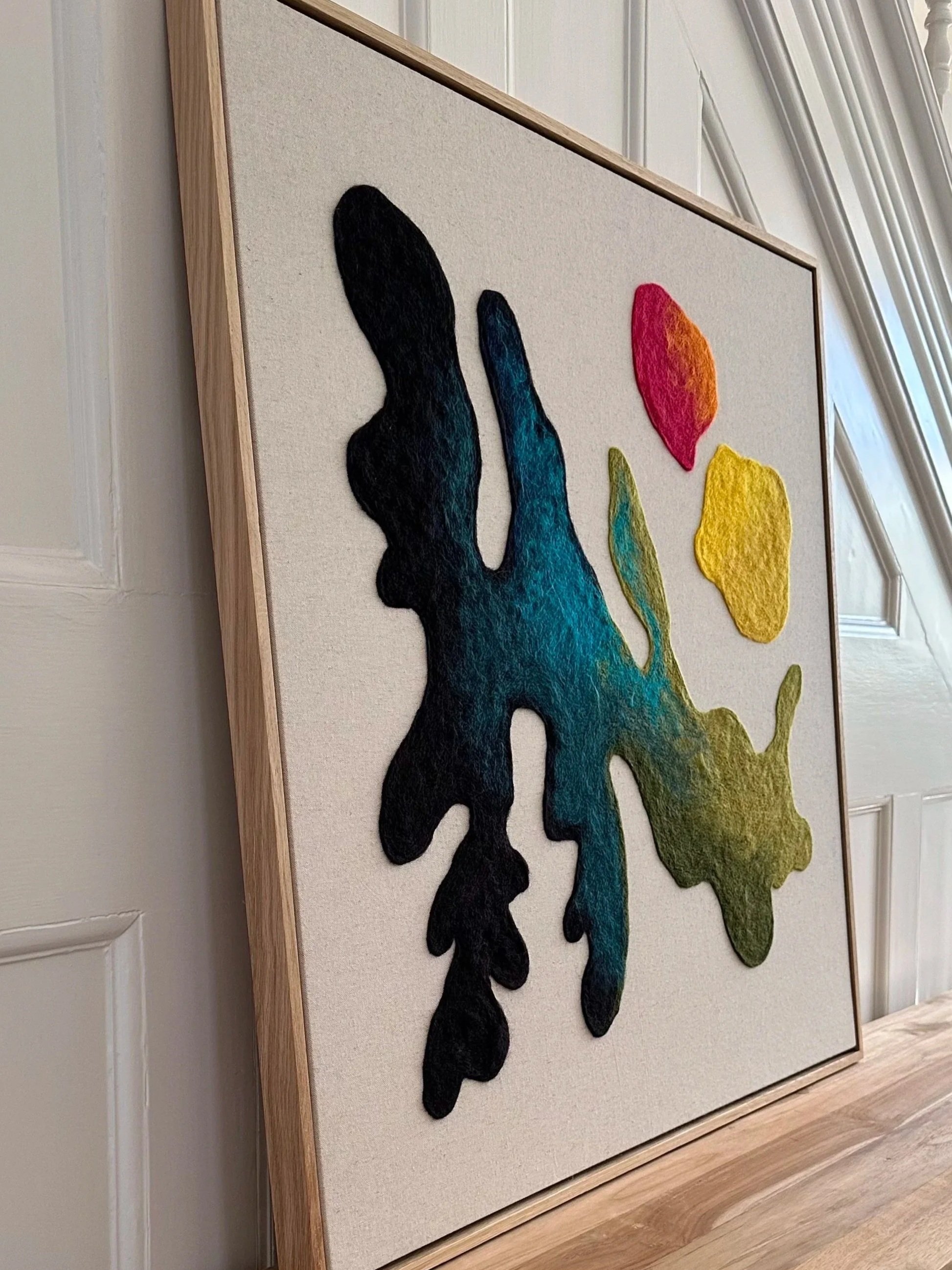 Colorful abstract artwork on canvas, featuring an organic black shape with hues of blue, green, and yellow, and three smaller patches of red, orange, and yellow, framed in light wood, hanging on a white paneled wall.