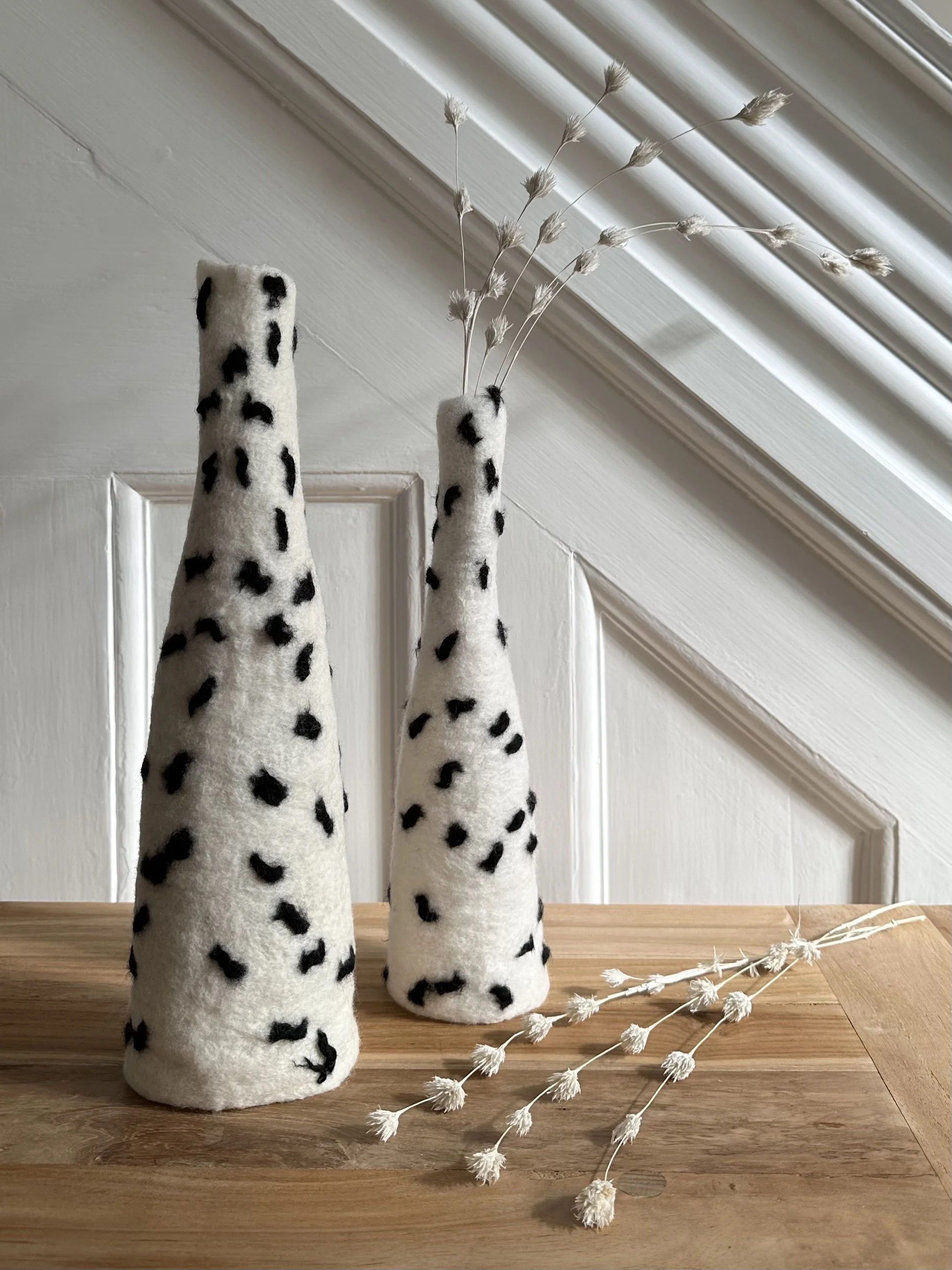 Two tall, cone-shaped vases with black and white spotted patterns, one with dried white flowers and the other empty, placed on a wooden surface near a white paneled wall.