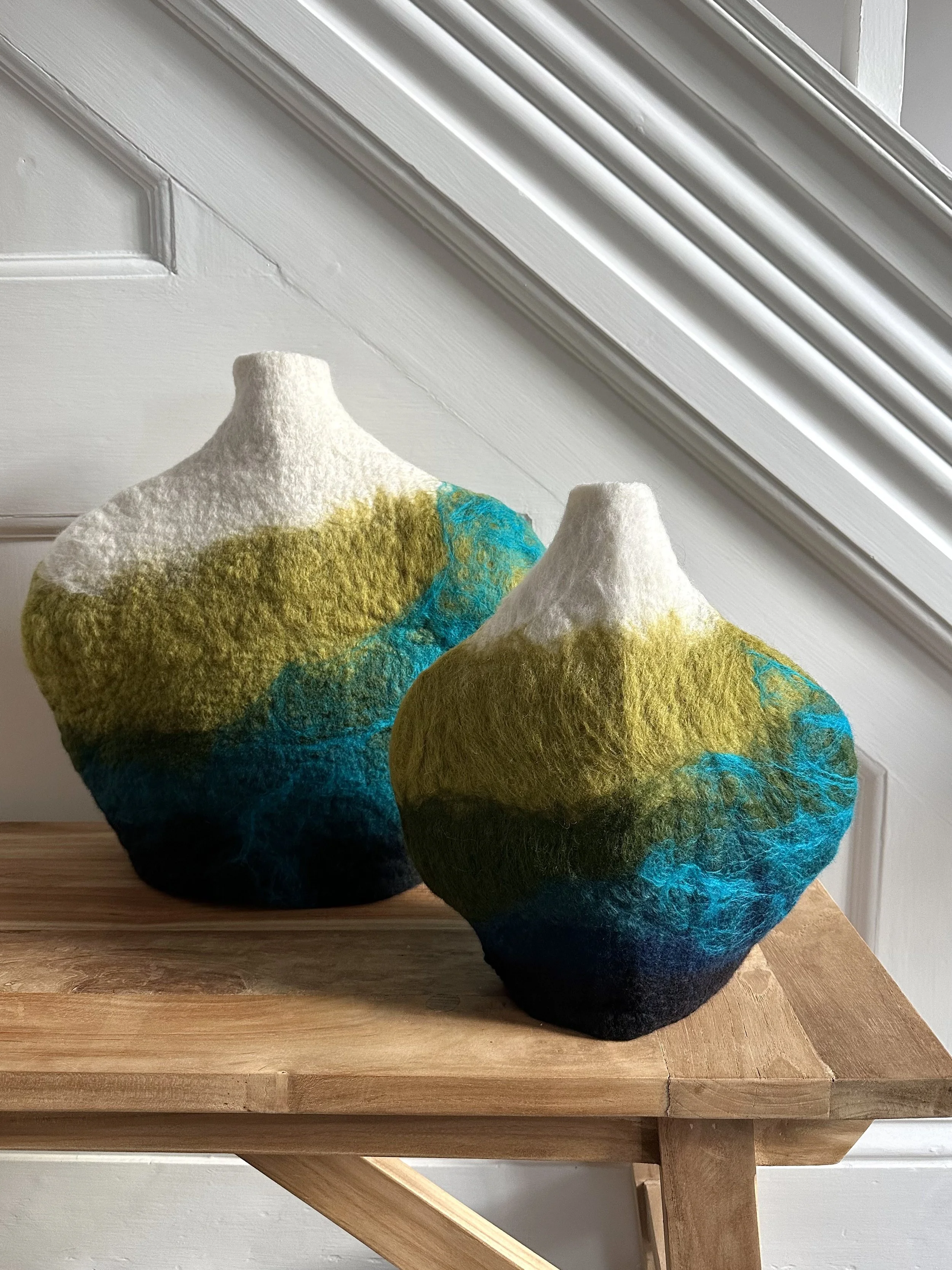 Two colorful felted vases with abstract landscape patterns on a wooden surface, against a white wall with crown molding.