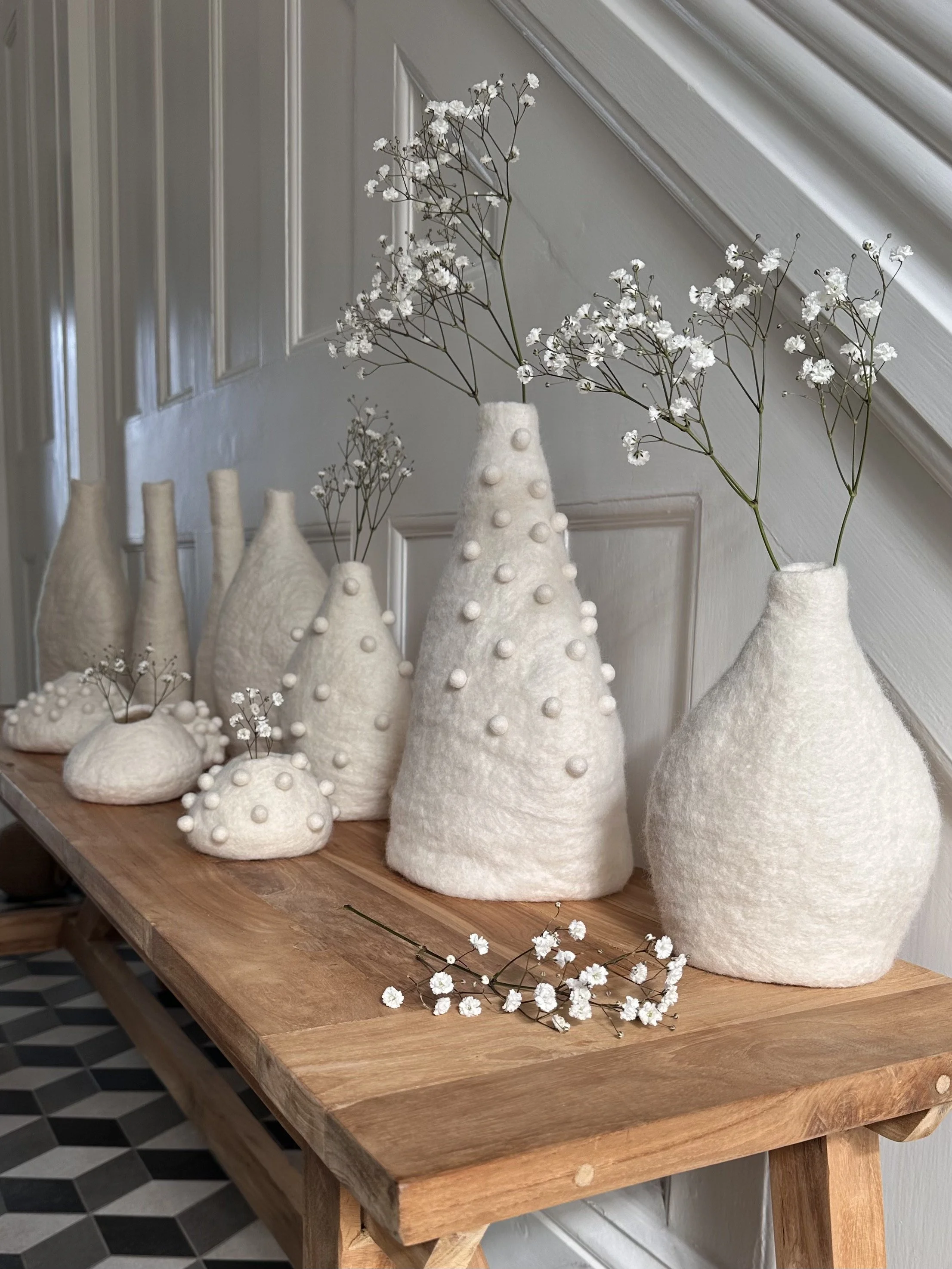 A wooden table decorated with white felt vases in various shapes and sizes, some with small white flowers and others with larger sprigs of baby's breath.