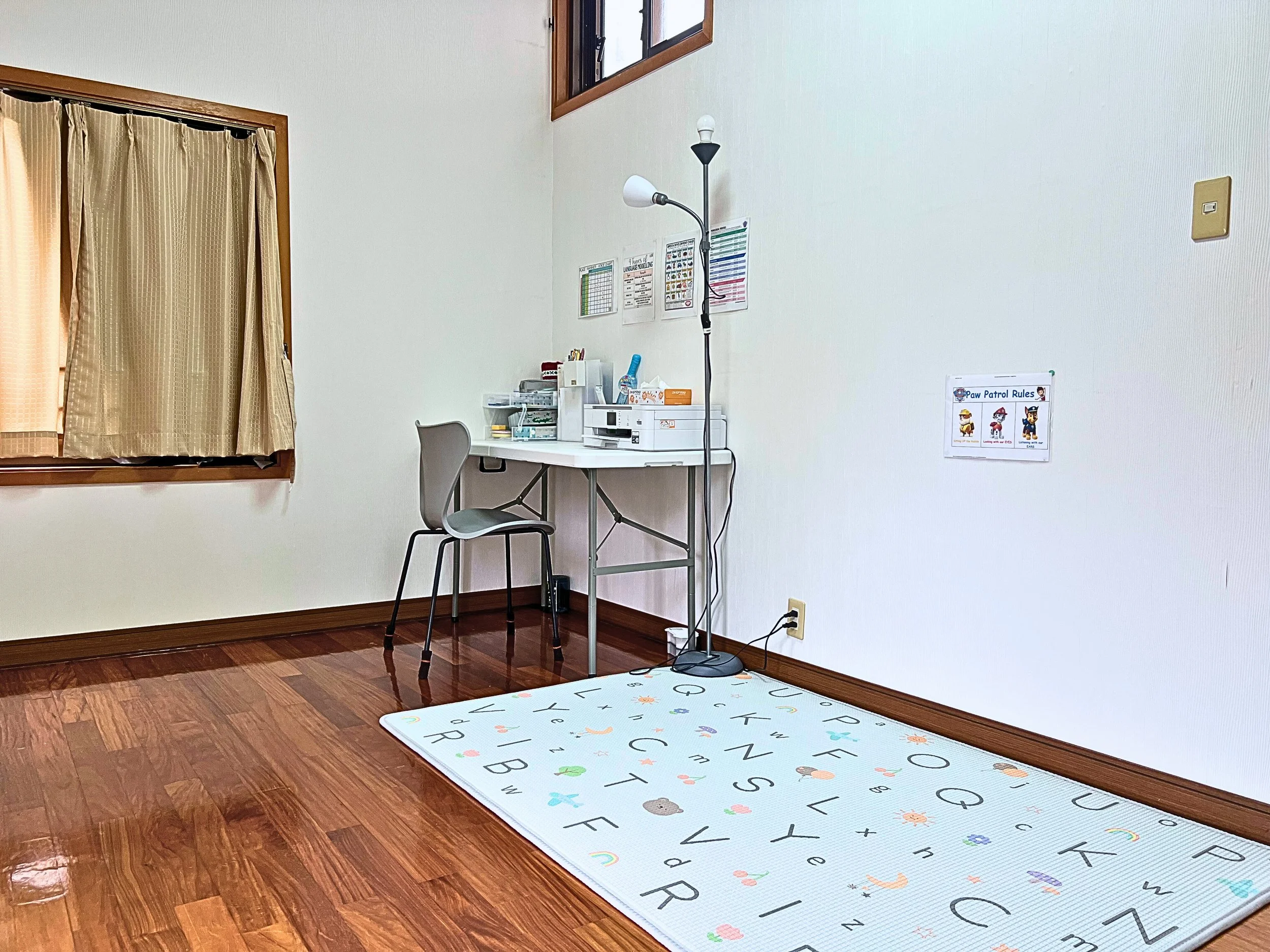 A small, organized workspace with a white desk, grey chair, floor lamp, and a colorful alphabet-themed rug on a shiny wooden floor, near a window with beige curtains.