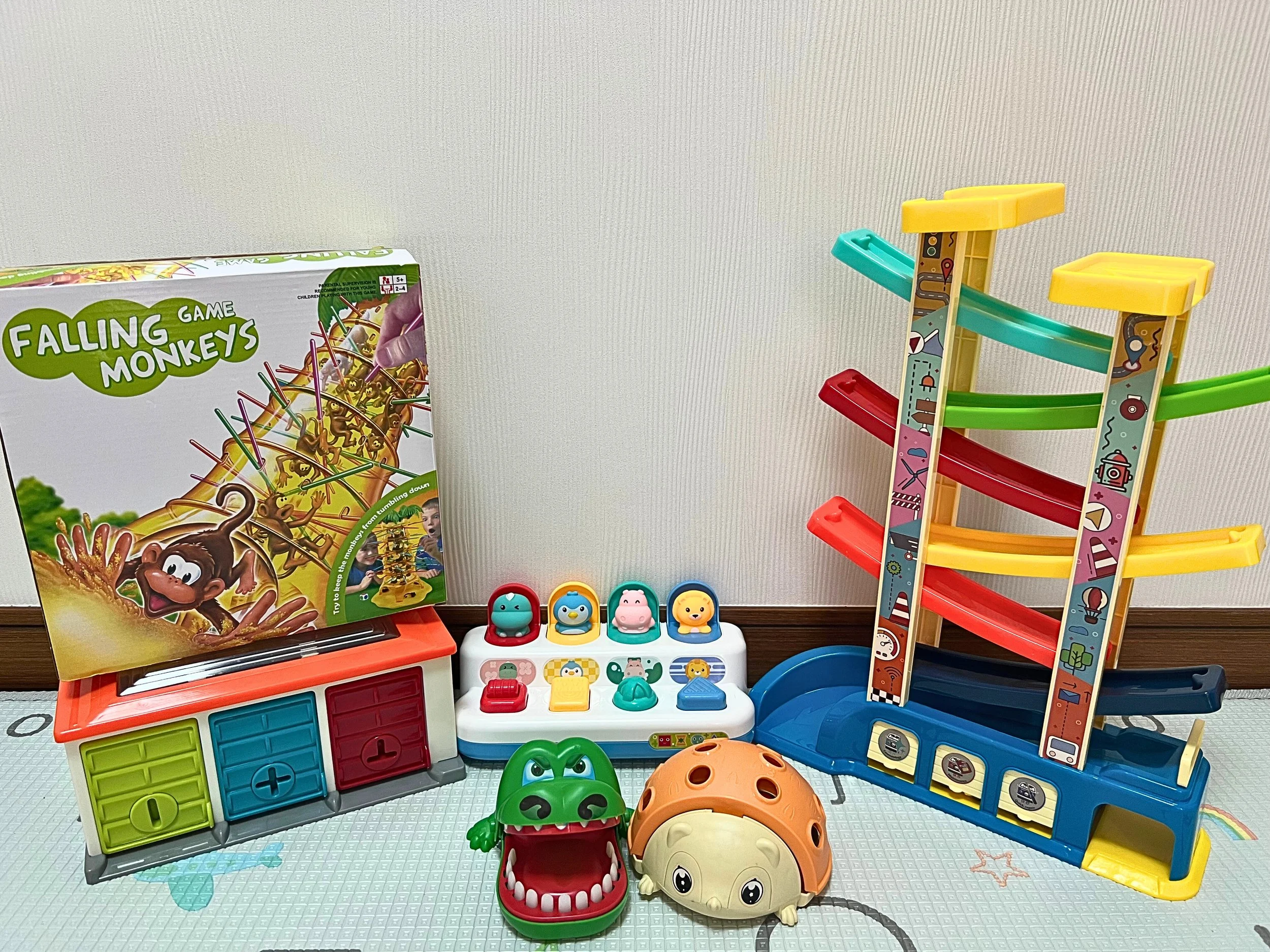 Children's toys including a colorful marble run, a monster-shaped toy, a ladybug toy, a game box titled 'Falling Monkeys', a toy with animal faces, and a small electronic toy, all arranged on a surface against a plain wall.