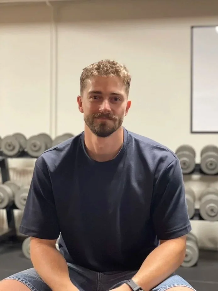 Ung mand med mørkt hår og skæg, sidder i et fitnesscenter med vægte i baggrunden.