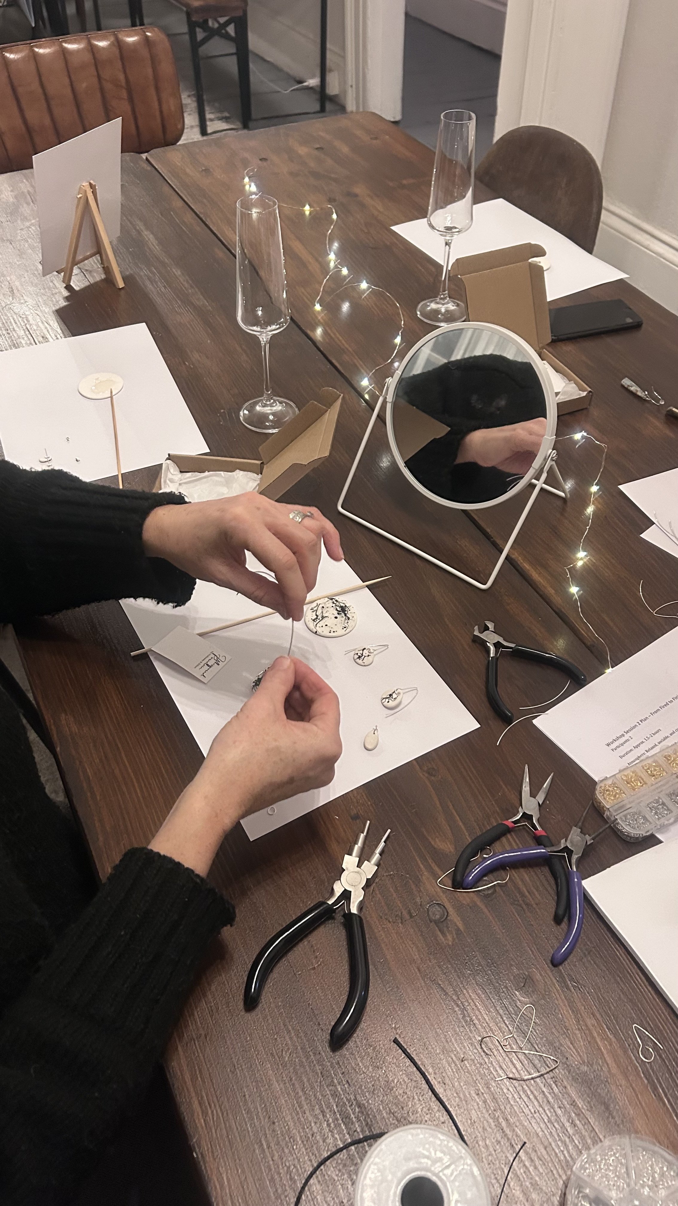 Person making jewelry at a wooden table with tools, a mirror, and jewelry pieces, in a cozy indoor setting.
