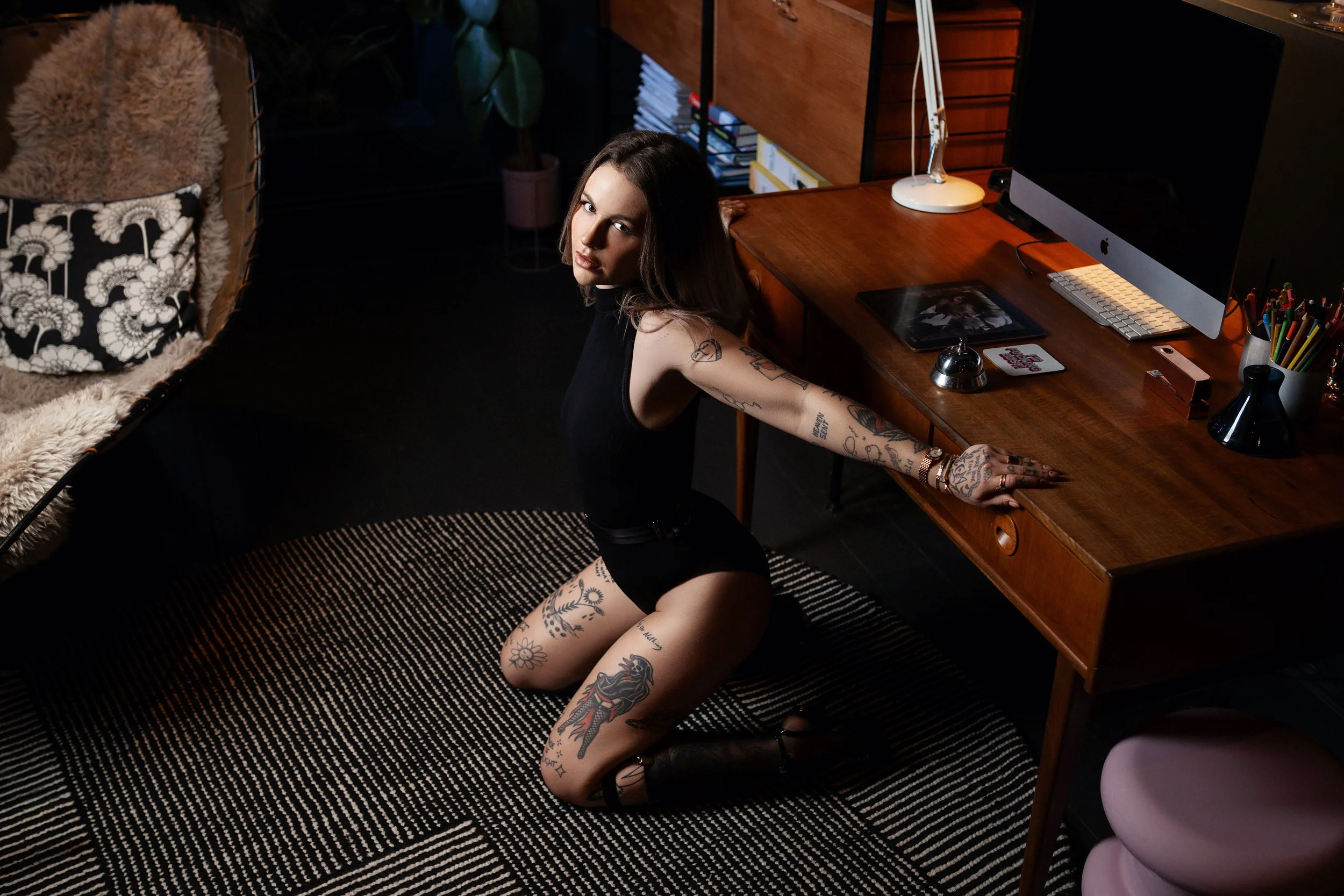 A woman with tattoos on her arms and legs kneeling on a black-and-white striped rug in front of a wooden desk with a computer, mouse, and various office supplies in a dimly lit room.