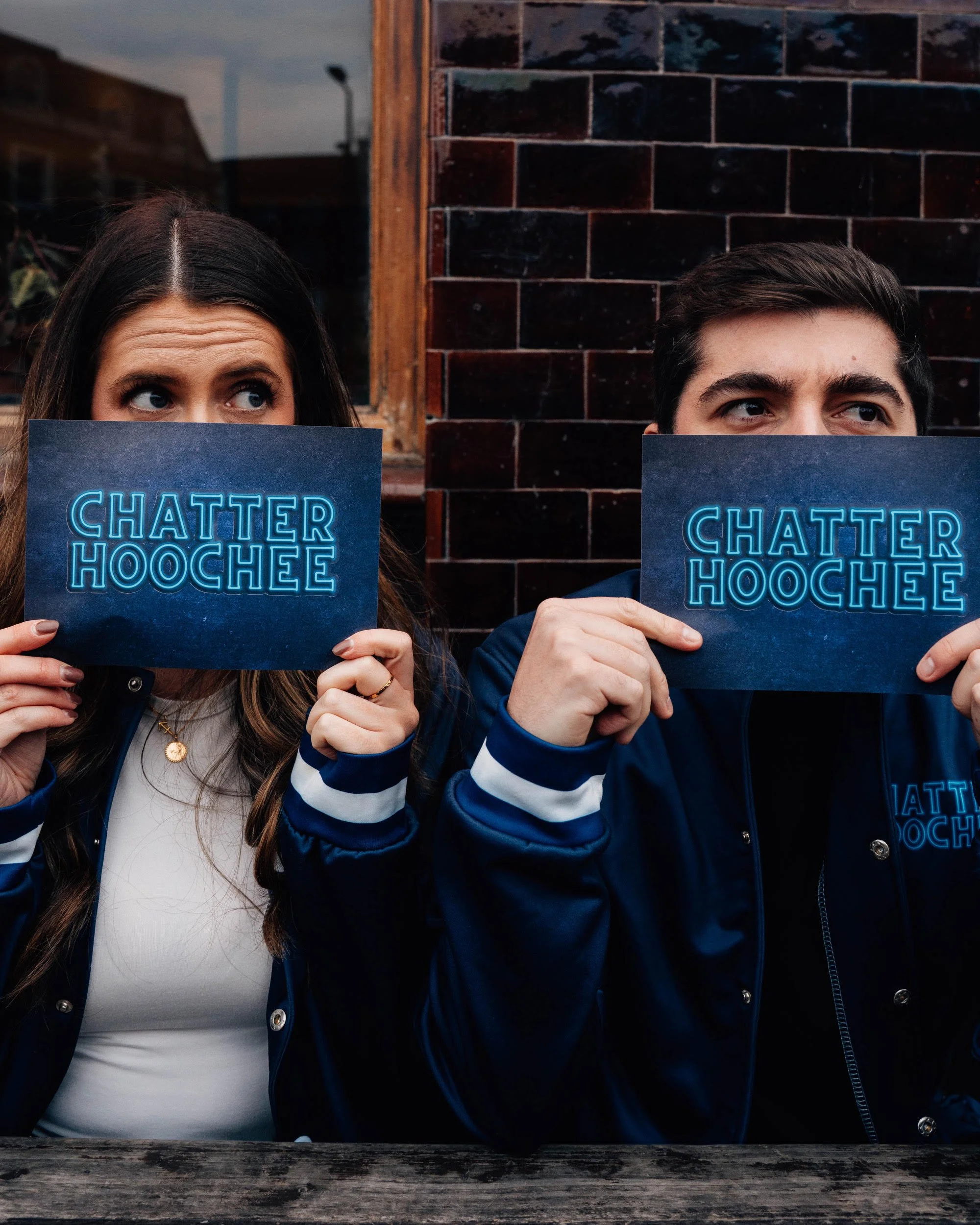 Two young adults, a woman with long brown hair and a man with short dark hair, holding blue signs that say 'Chatter Hoochee' and peering over them with curious expressions. They are sitting at a wooden table outside in front of a brick wall.