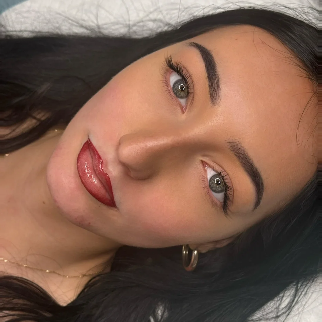 Close-up of a woman's face showing her with black hair, brown eyes, red lipstick, and a small hoop earring.