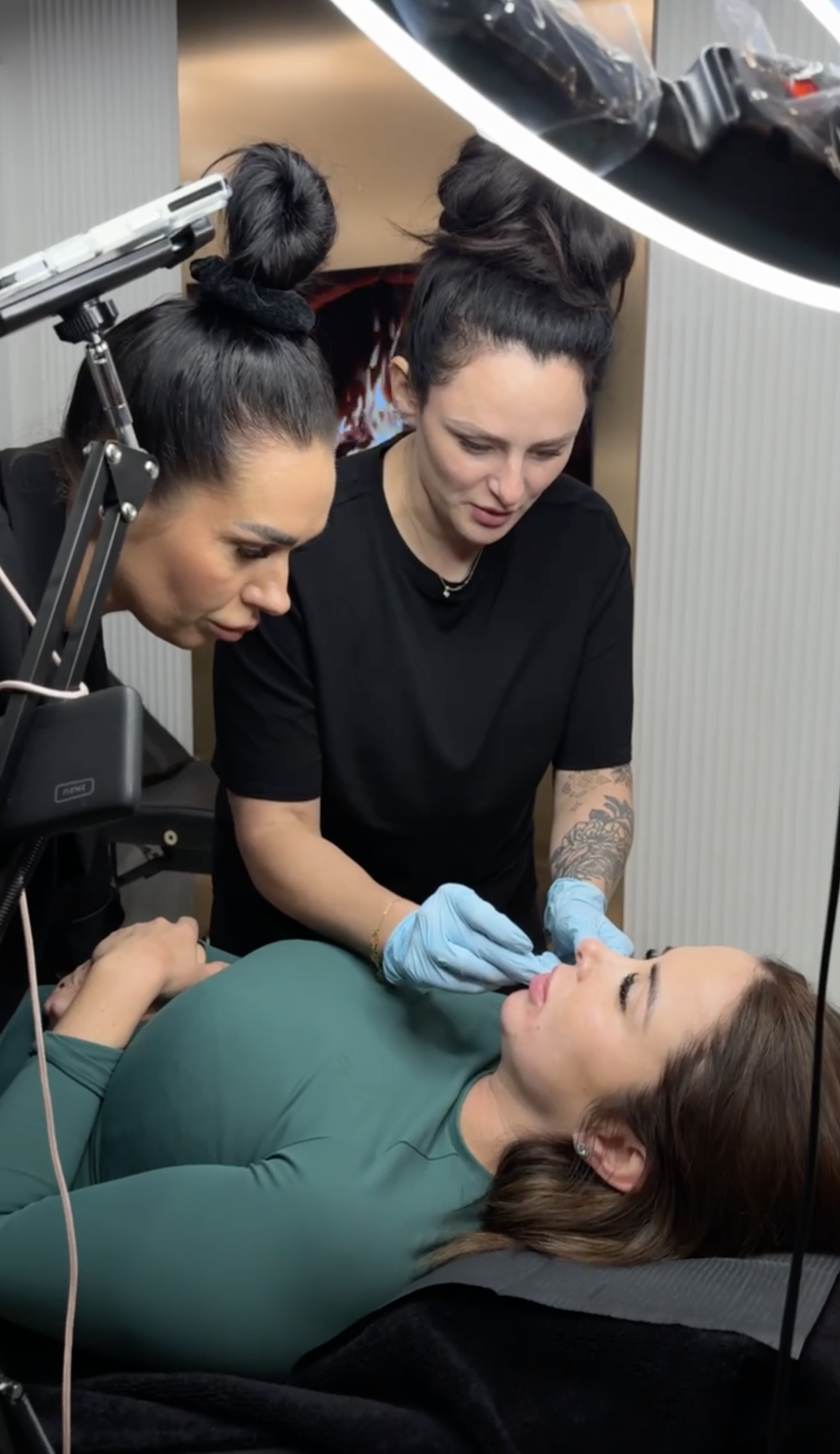 A woman lying on a treatment bed as two professionals perform a cosmetic procedure, using tools and wearing gloves, in a clinical setting.