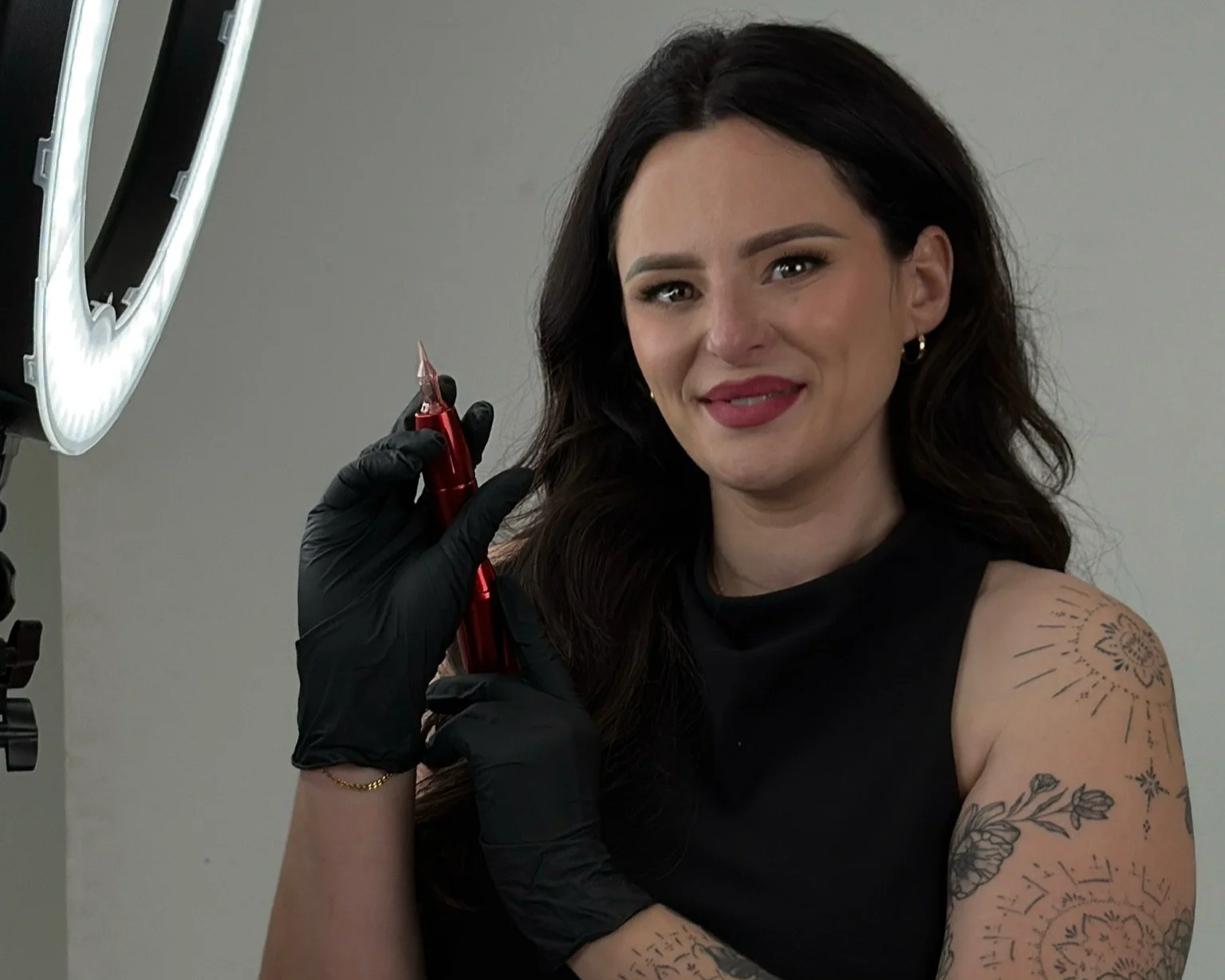 Woman with dark hair and tattoos getting a tattoo on her shoulder, holding an electric tattoo pen, wearing black gloves and a black top.