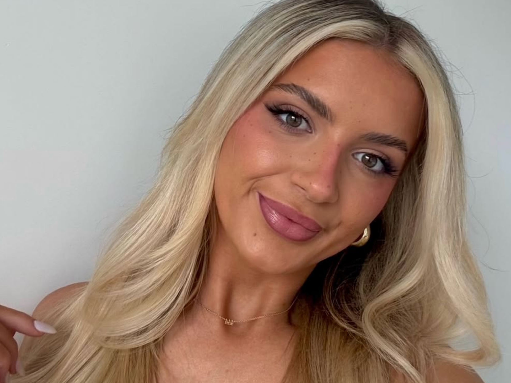 A young woman with long, wavy blonde hair, wearing makeup with pink lipstick and eye makeup, smiling at the camera, against a gray background.