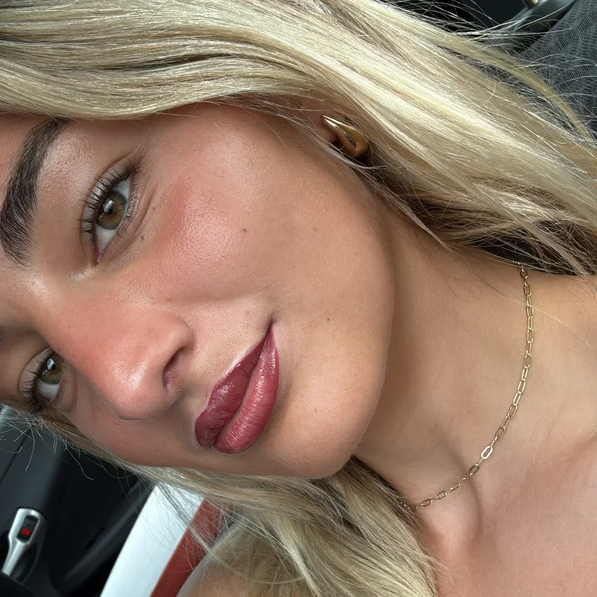 A close-up of a woman's face with blonde hair, wearing gold jewelry, and a black top, lying in a car.