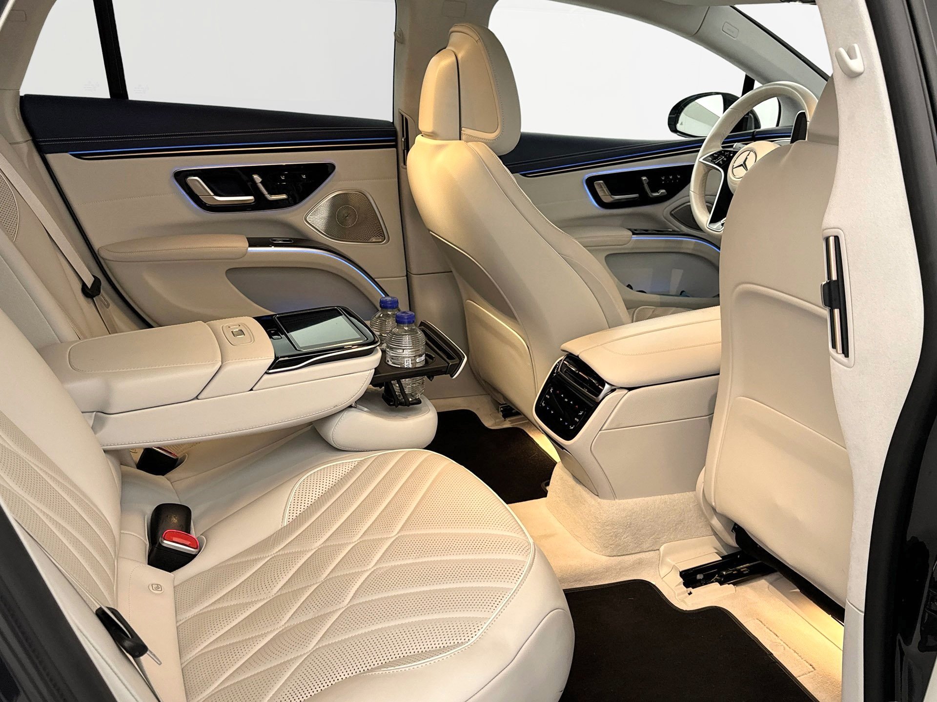 Interior of a luxury car with beige leather seats, armrests, and controls, including water bottles in the rear cup holder and climate vents.