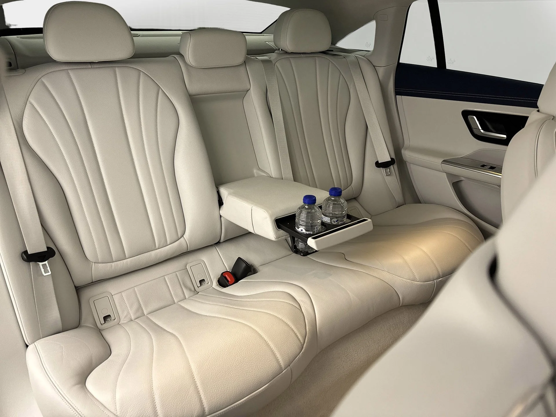 The interior of a vehicle's back seat showing beige leather seats, a fold-down armrest with two water bottles, and a section of the door with a handle and window controls.
