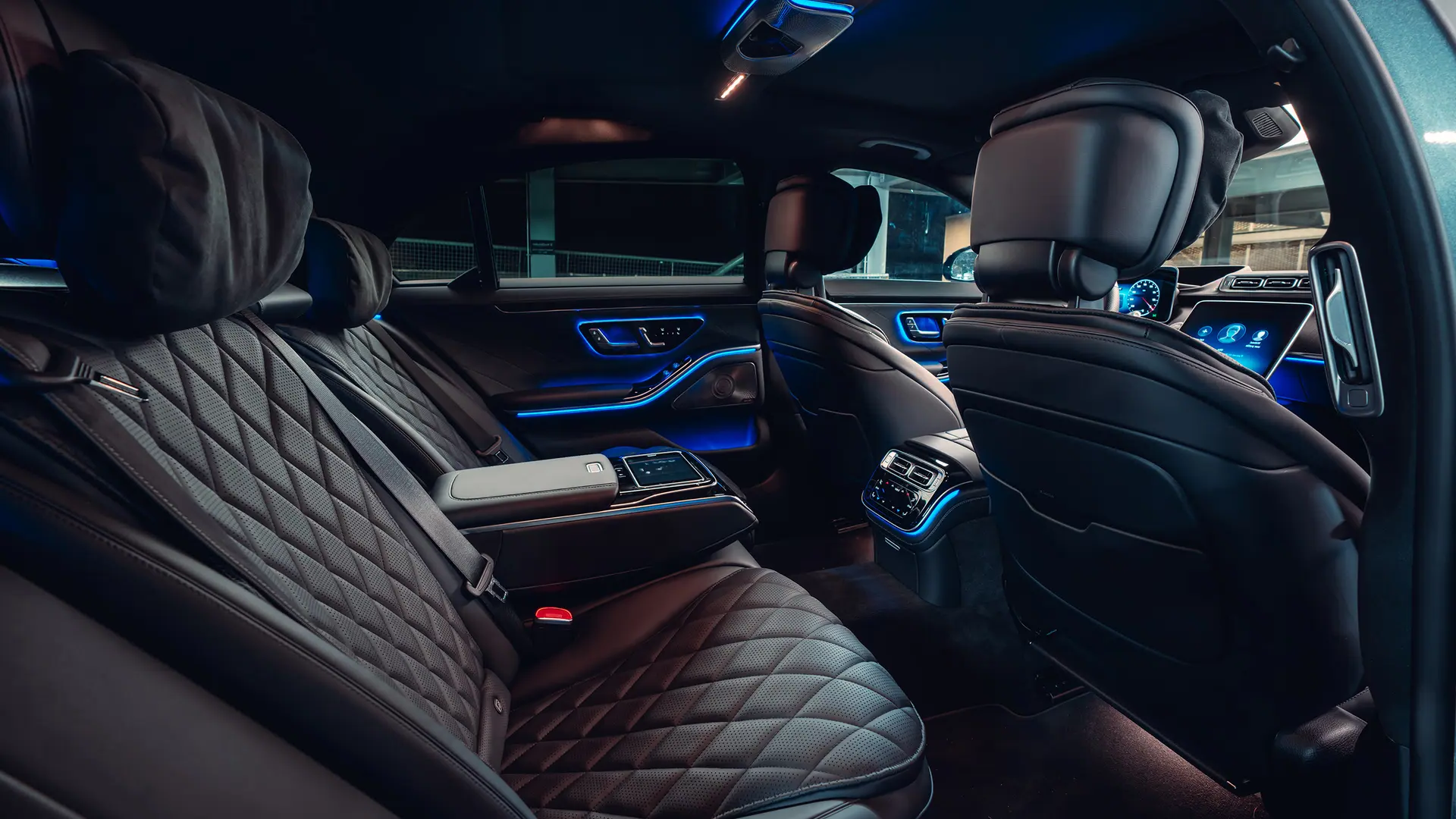 The interior of a luxury car showing quilted leather seats with adjustable headrests, illuminated ambient lighting along the doors and dashboard, central control console with a touch display, and a digital instrument cluster in the front.