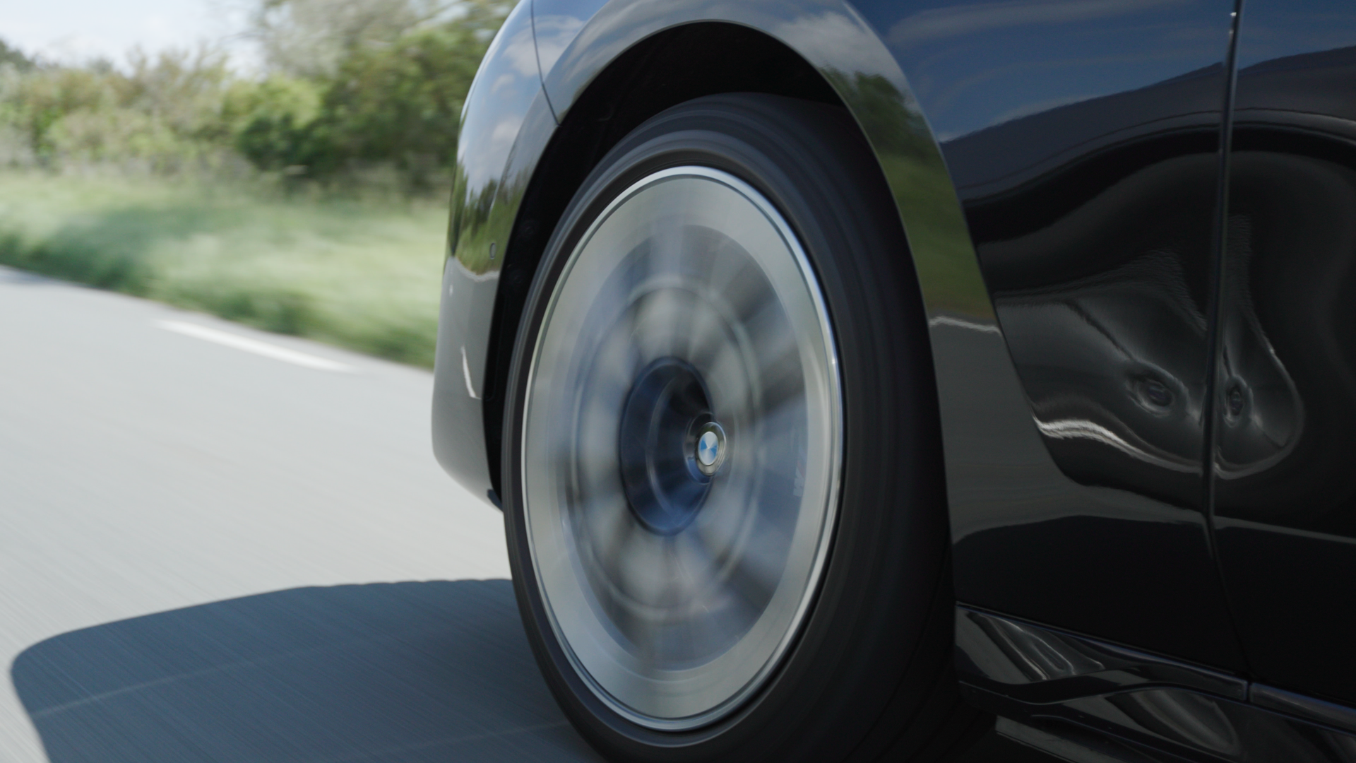 Close-up of a moving black car's front wheel on a road, with a blurred background of green trees and sky, and a visible reflection on the car's body.