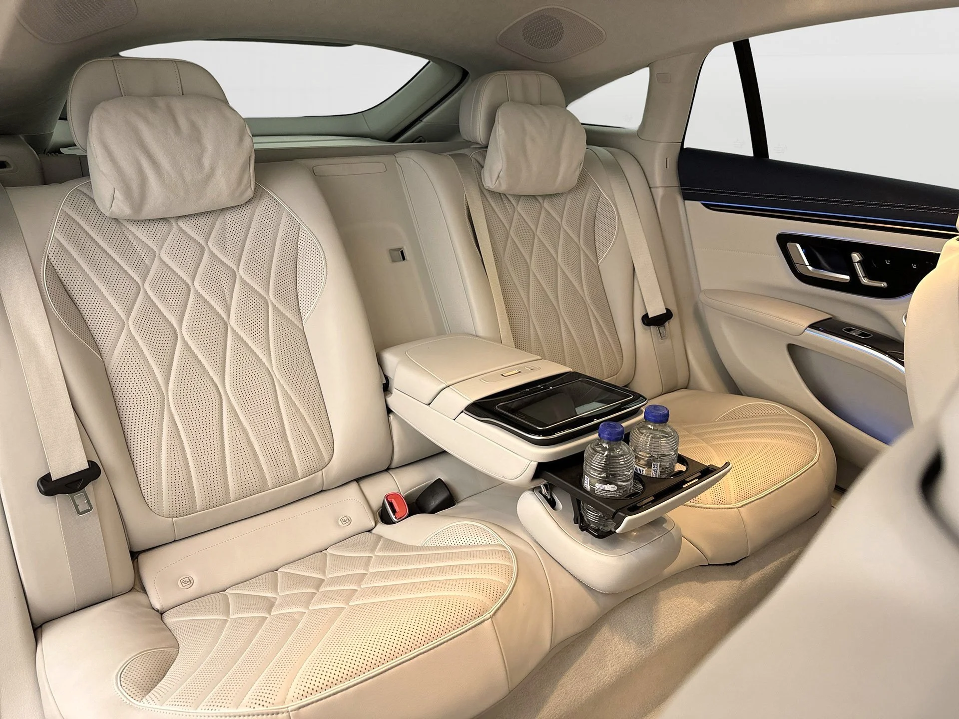 The backseat of a luxury car with beige leather seats, headrests, and built-in controls. Two water bottles are in a fold-out tray and the interior door panel is visible.