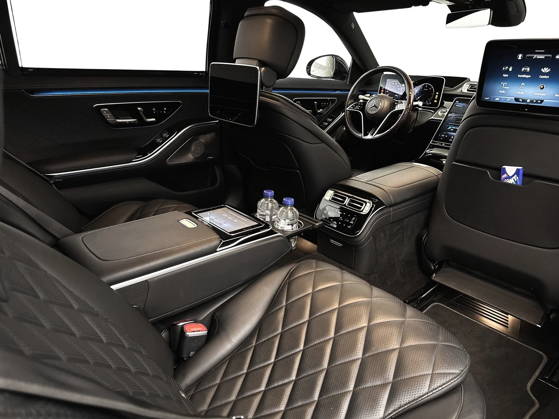 The interior of a luxury car with black leather seats, quilted pattern on the seats, a center armrest with a tablet, two water bottles, digital screens on the dashboard and back of the front seats, and modern controls.