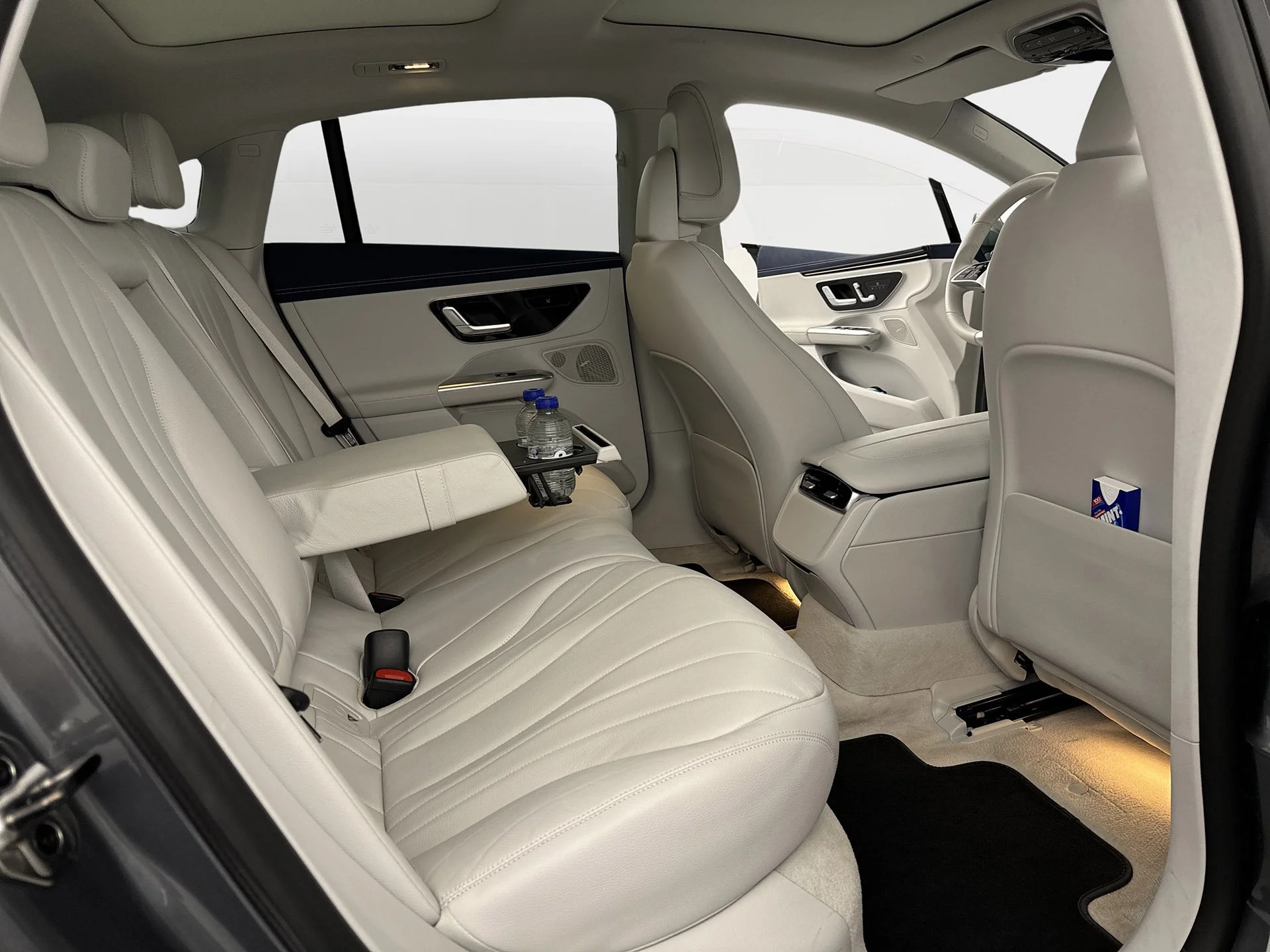 The interior of a luxury vehicle with beige leather seats, including the back seats with a fold-down armrest that has a water bottle holder, and the front seats with a modern dashboard and controls.