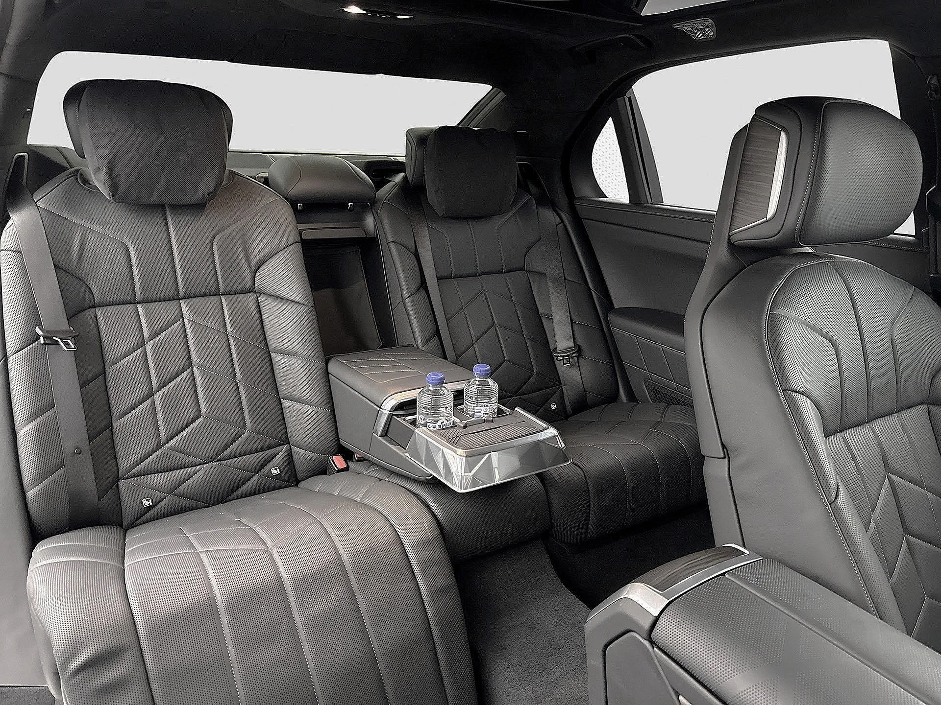 The interior of a luxury vehicle showing black leather seats with diamond stitching, a fold-down center console with two bottled waters, and a spacious back seat area with headrests, side windows, and sleek design details.