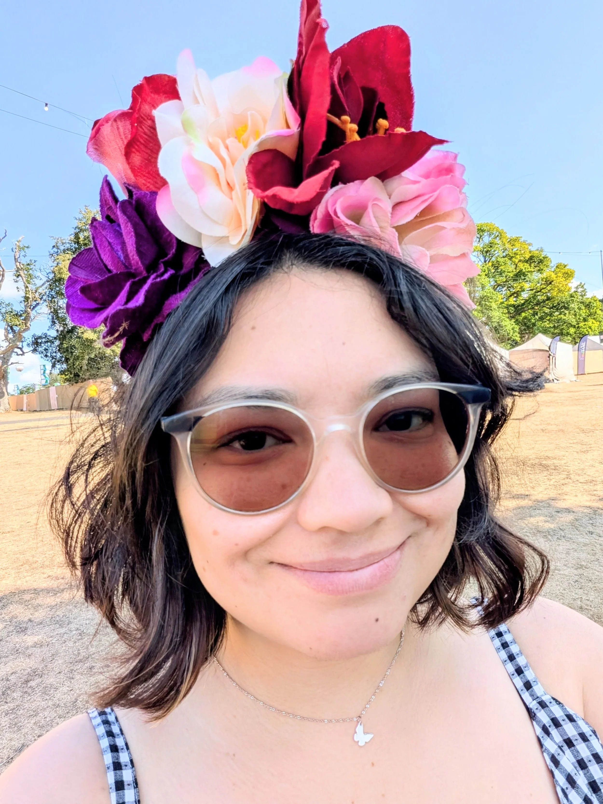 photo of me smiling in the sun wearina butterfly nexklace, sunglasses and a big colourful flower crown in a field