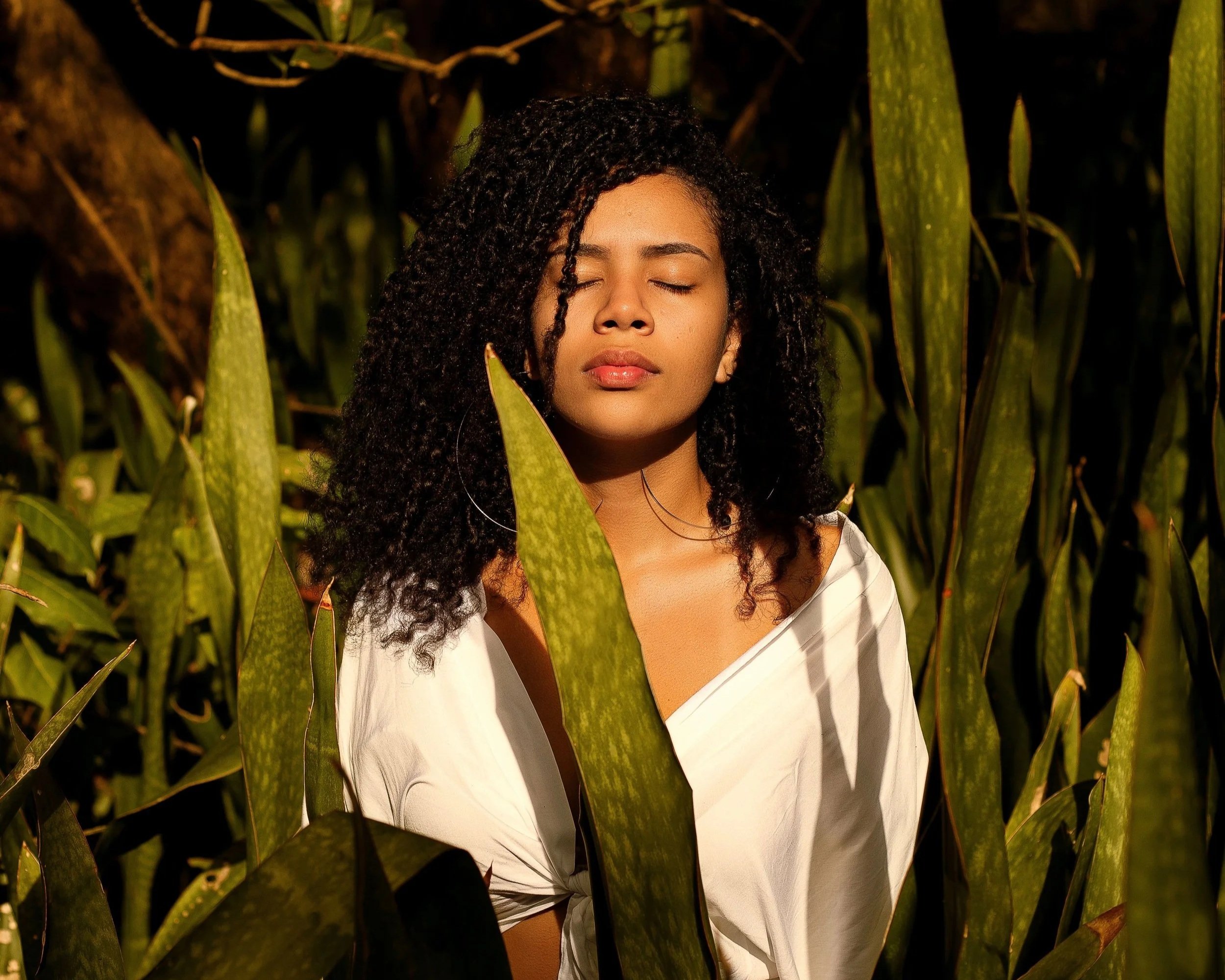 A woman of colour stands surrounded by nature with the sun on her face.