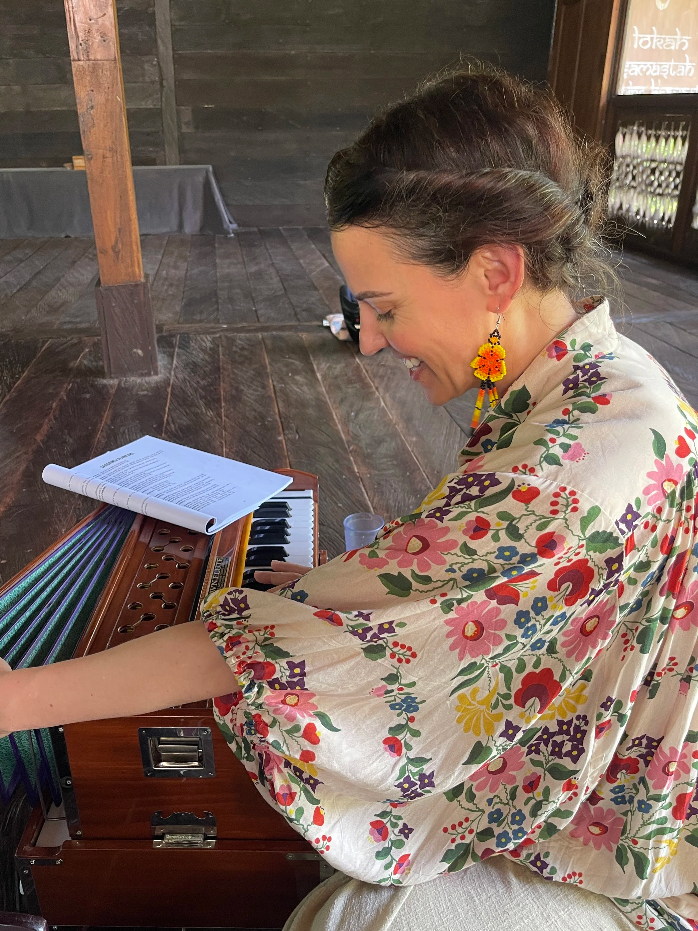 A woman with dark hair styled in a braided crown, wearing a colorful floral dress and a yellow flower-shaped earring, sitting at a wooden table playing a small pink keyboard. There are papers, possibly sheet music, on the keyboard.