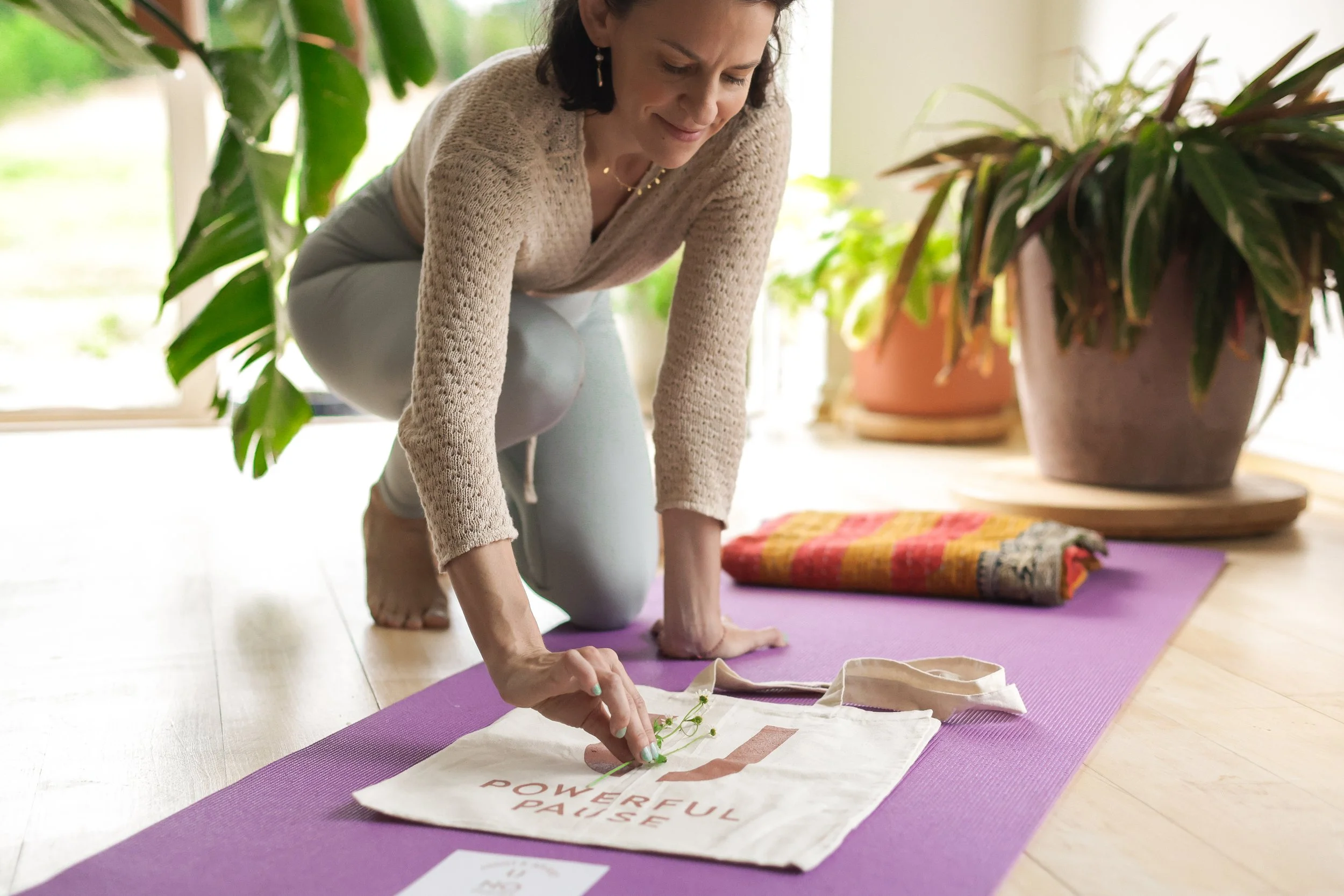 Woman in a beige sweater practicing yoga on a purple mat indoors, with green plants and natural light in the background.