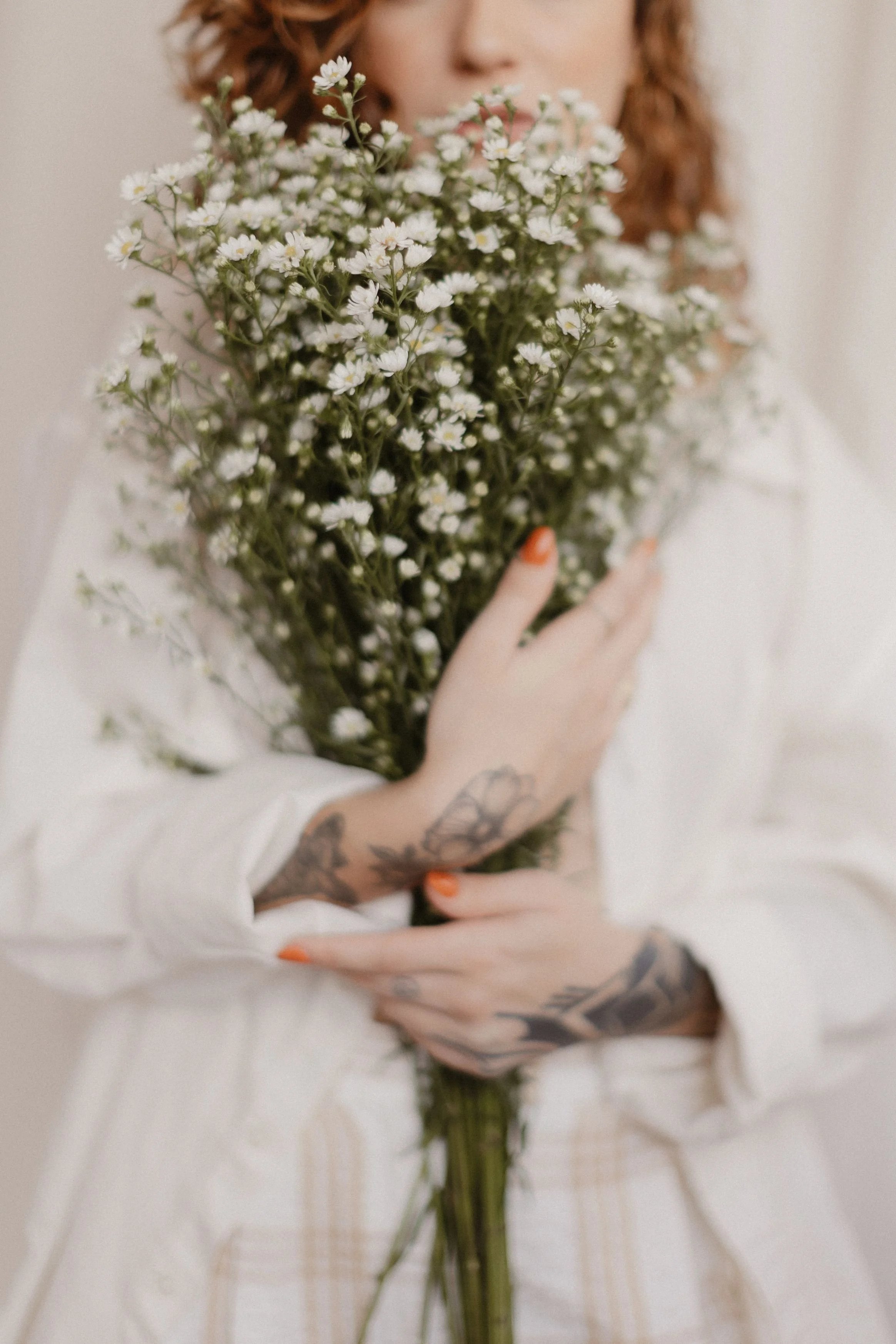 Person with tattoos on arms and hands, holding a bouquet of small white flowers with green stems, wearing a white long-sleeved shirt, against a neutral background.