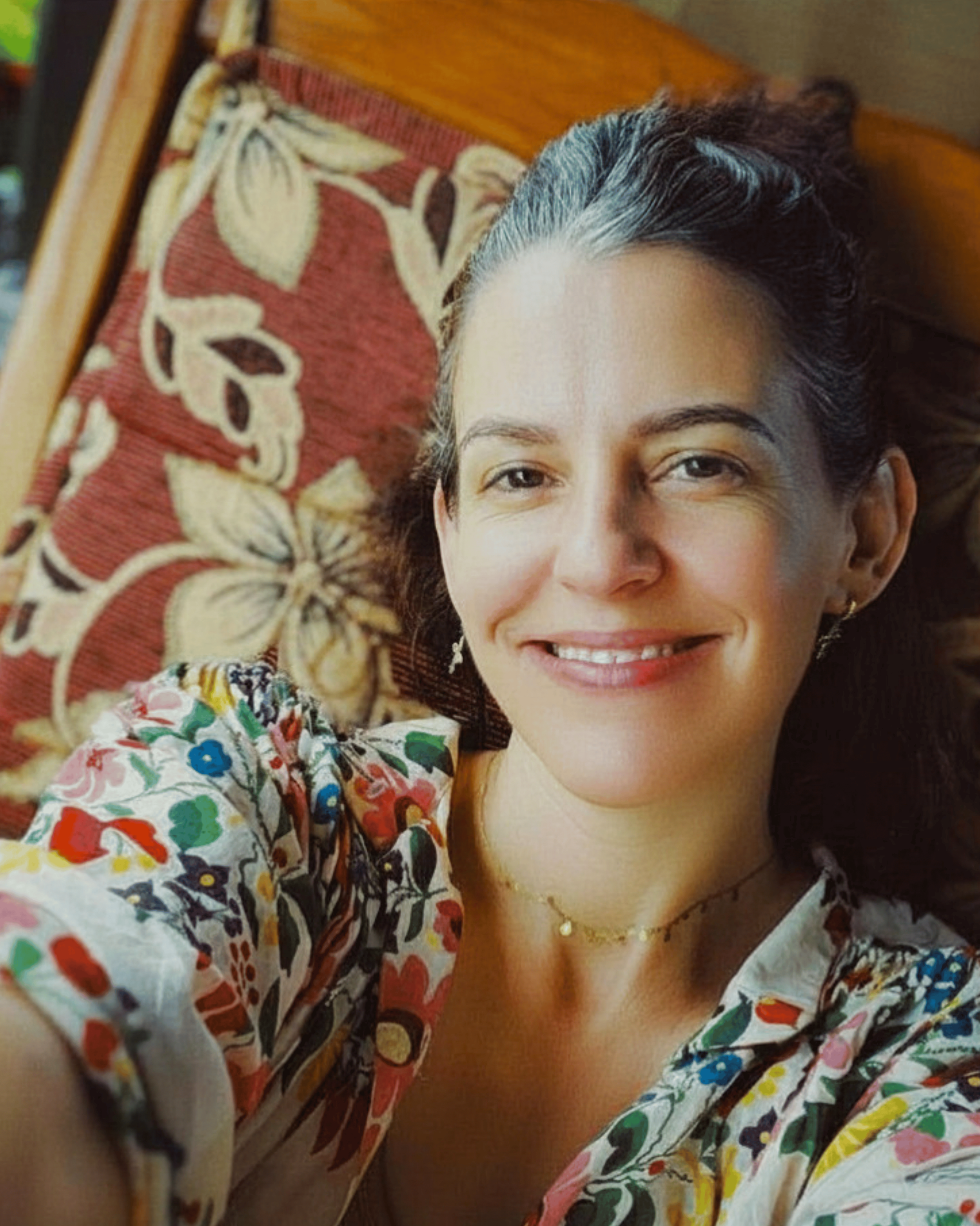 A woman with dark hair and a bright, colorful floral dress, lying on a patterned cushioned surface, smiling.