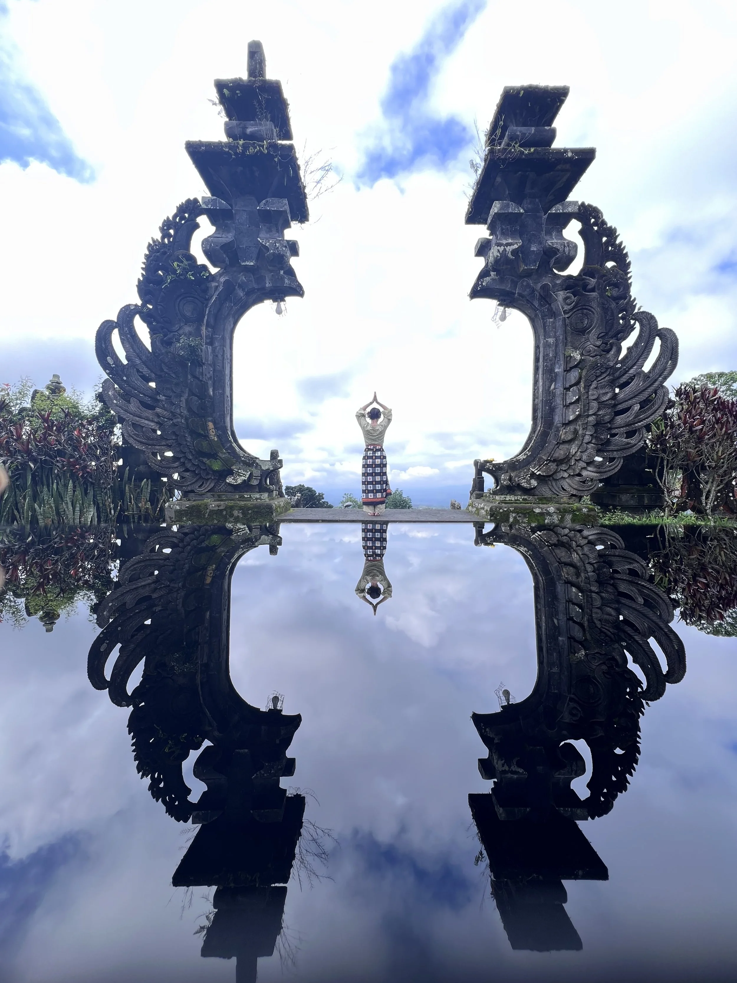 Traditional Balinese gateway with intricate carvings, reflecting in a pool of water with a cloudy sky background, and a woman standing with her hands in a prayer pose in the center.