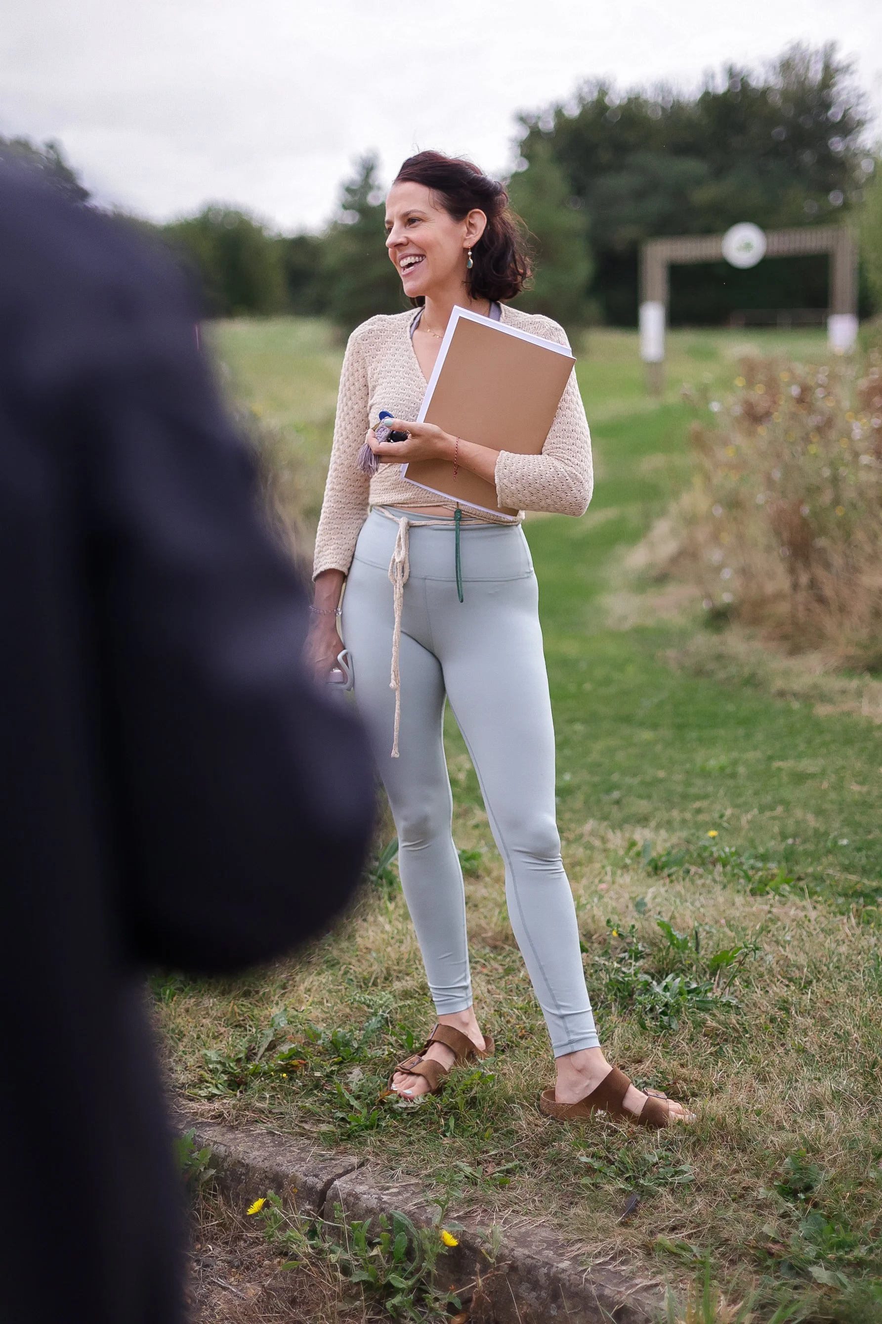 A woman with dark hair, wearing a crochet sweater, light-colored pants, and brown sandals, is smiling and talking outdoors, holding a notebook or folder. There is a blurred figure in the foreground and a park entrance in the background.
