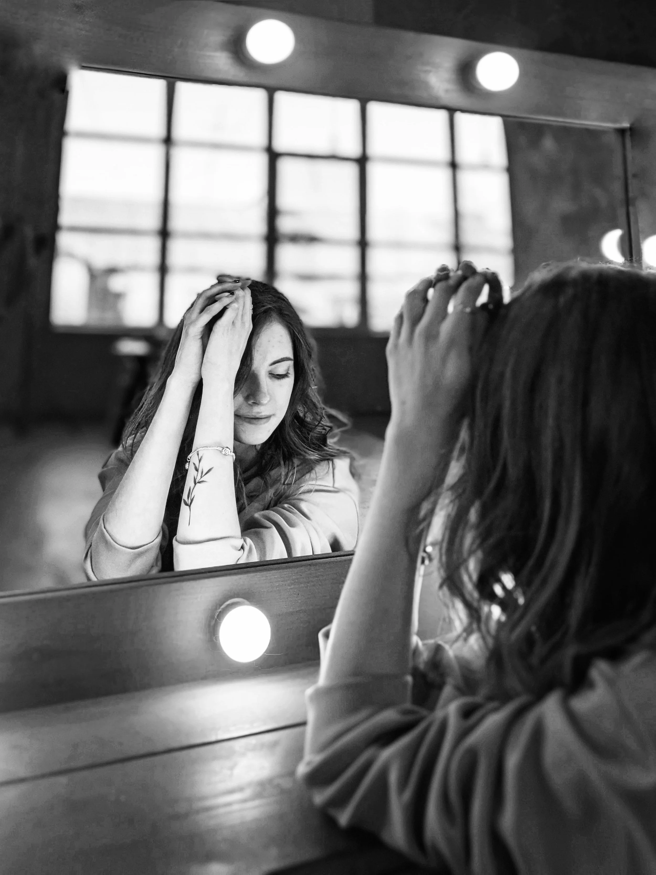 A woman with long hair and a floral patterned blouse, sitting at a vanity table, looking into a mirror with lights around the frame, adjusting her hair with her hands, in a black and white photo.