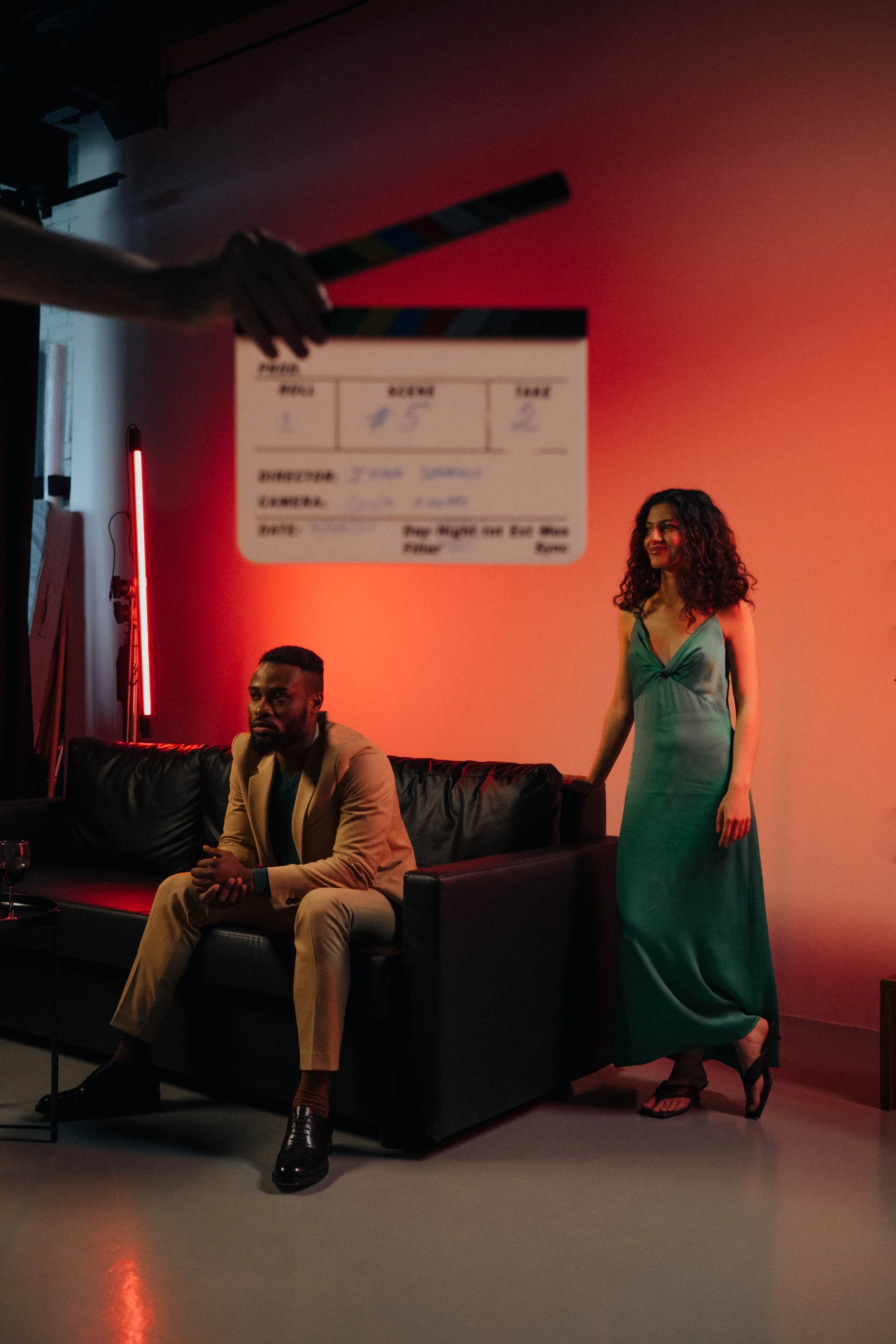 A studio scene with a woman in a long green dress standing next to a man in a tan suit sitting on a black couch. The woman has curly hair and is smiling, the man has short hair and a beard. There is a clapperboard in the foreground, slightly out of focus. The background is lit with red lighting, and a neon light is visible on the left side.