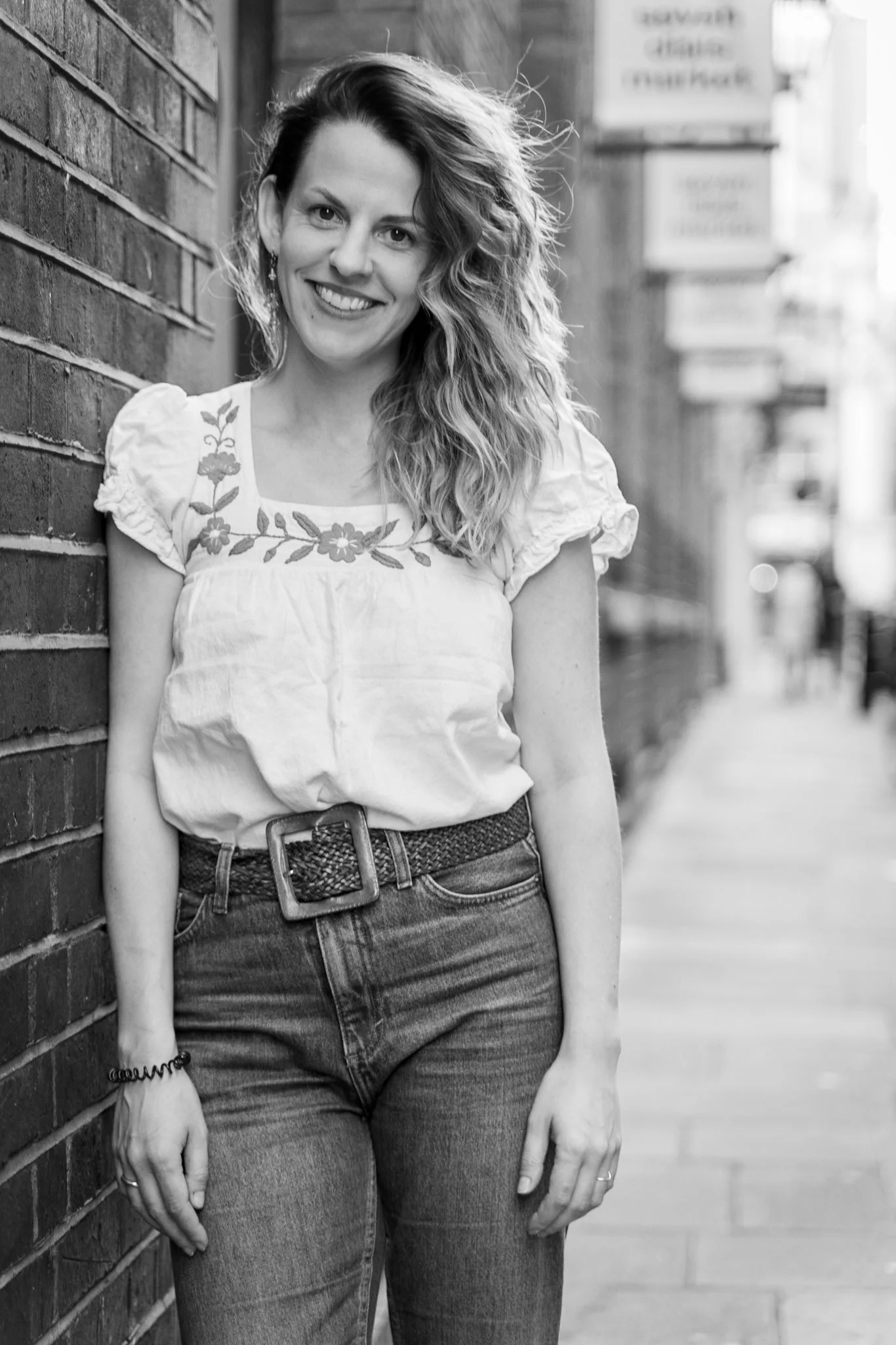 A smiling woman with curly hair standing outdoors, leaning against a brick wall in an urban setting.