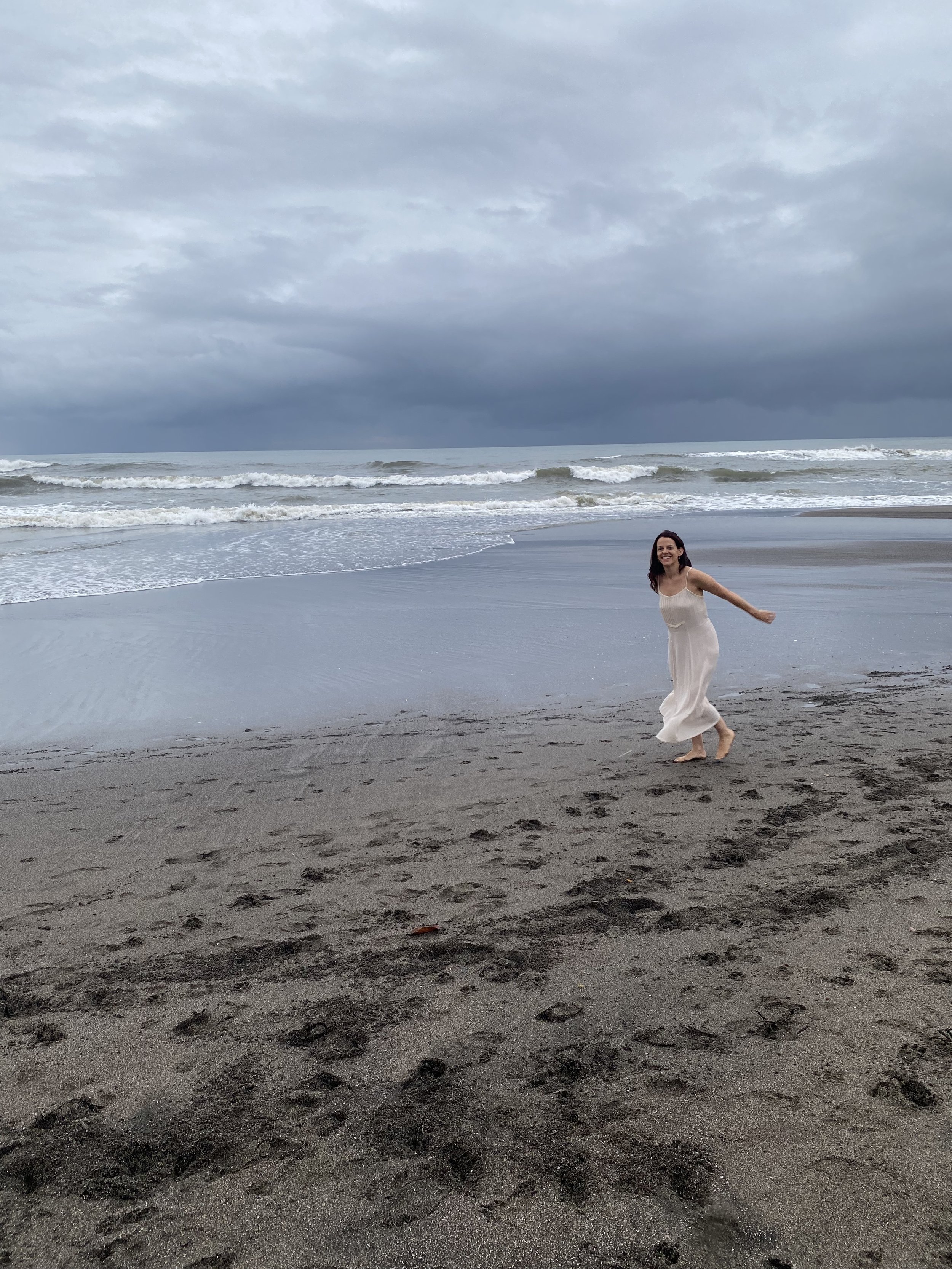 A woman in a white dress dancing on a sandy beach with dark clouds and ocean waves in the background.