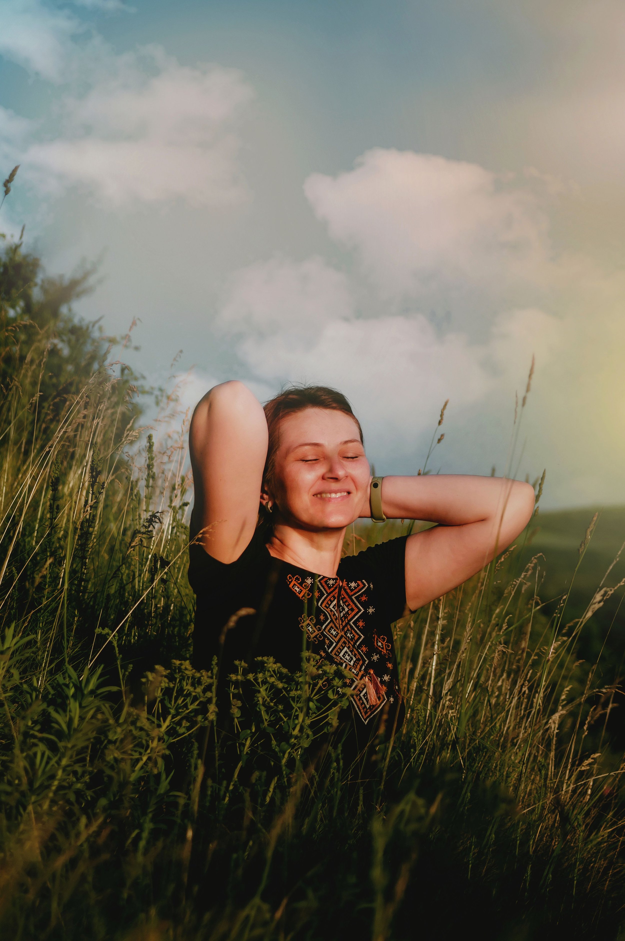 A smiling woman with closed eyes relaxing in a field of tall grass, with a sky filled with clouds behind her.
