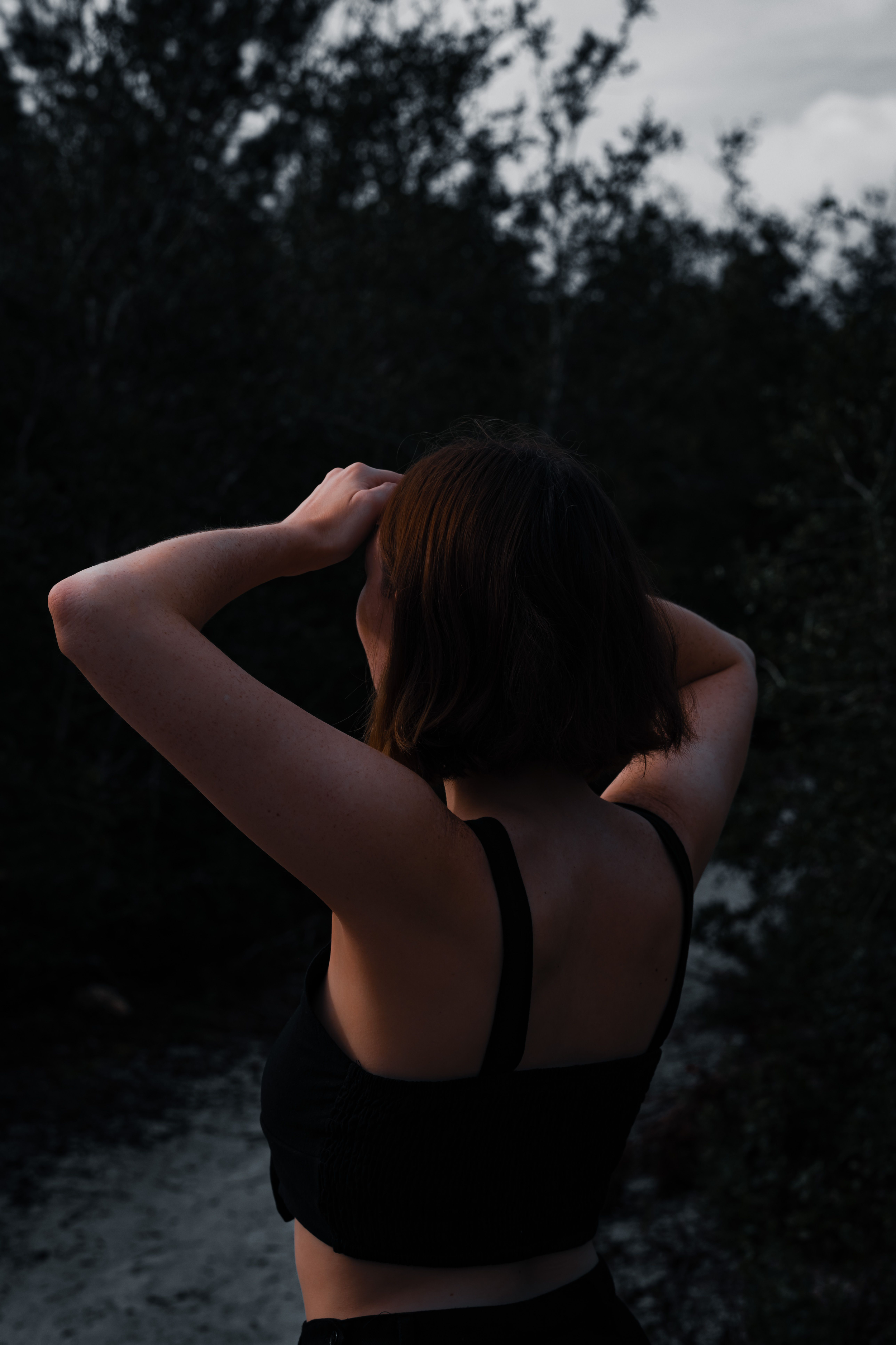A woman with short red hair and fair skin, wearing a black strappy top, standing outdoors in a natural setting with trees and a cloudy sky in the background. She has her hands raised to her head, and the photo is taken during twilight or early evening, with low light.