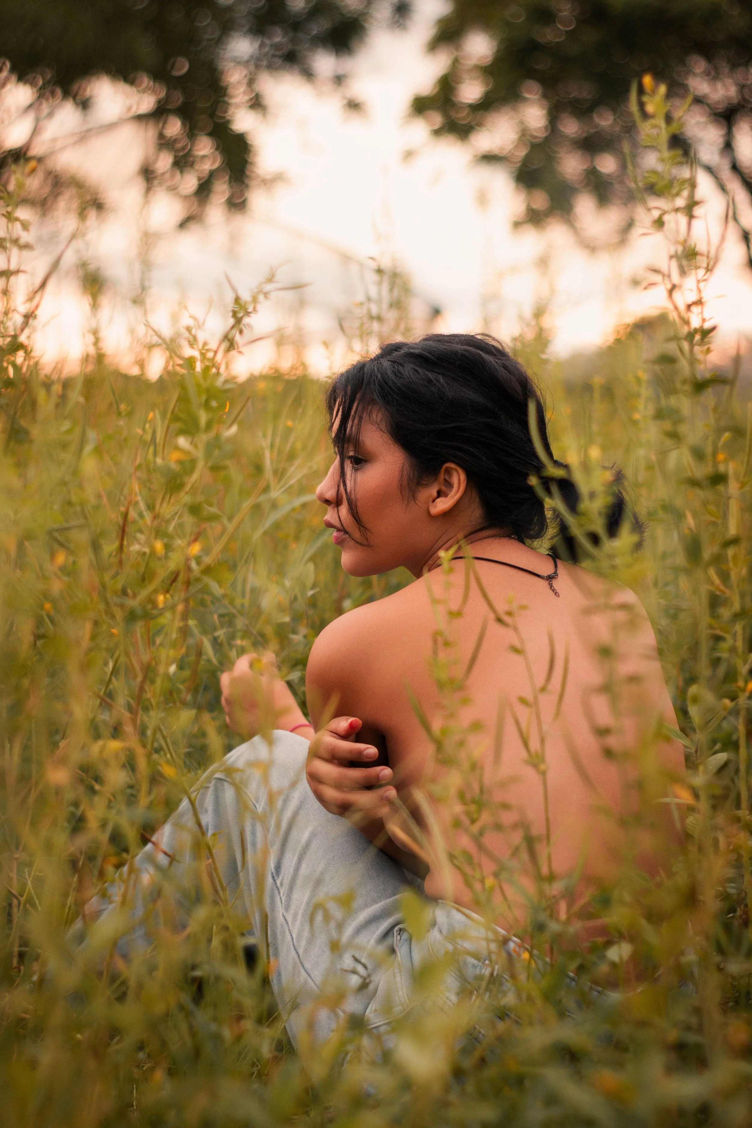 A woman with dark hair and a necklace sitting in a field of plants during sunset, with her head turned to the side and her eyes closed.