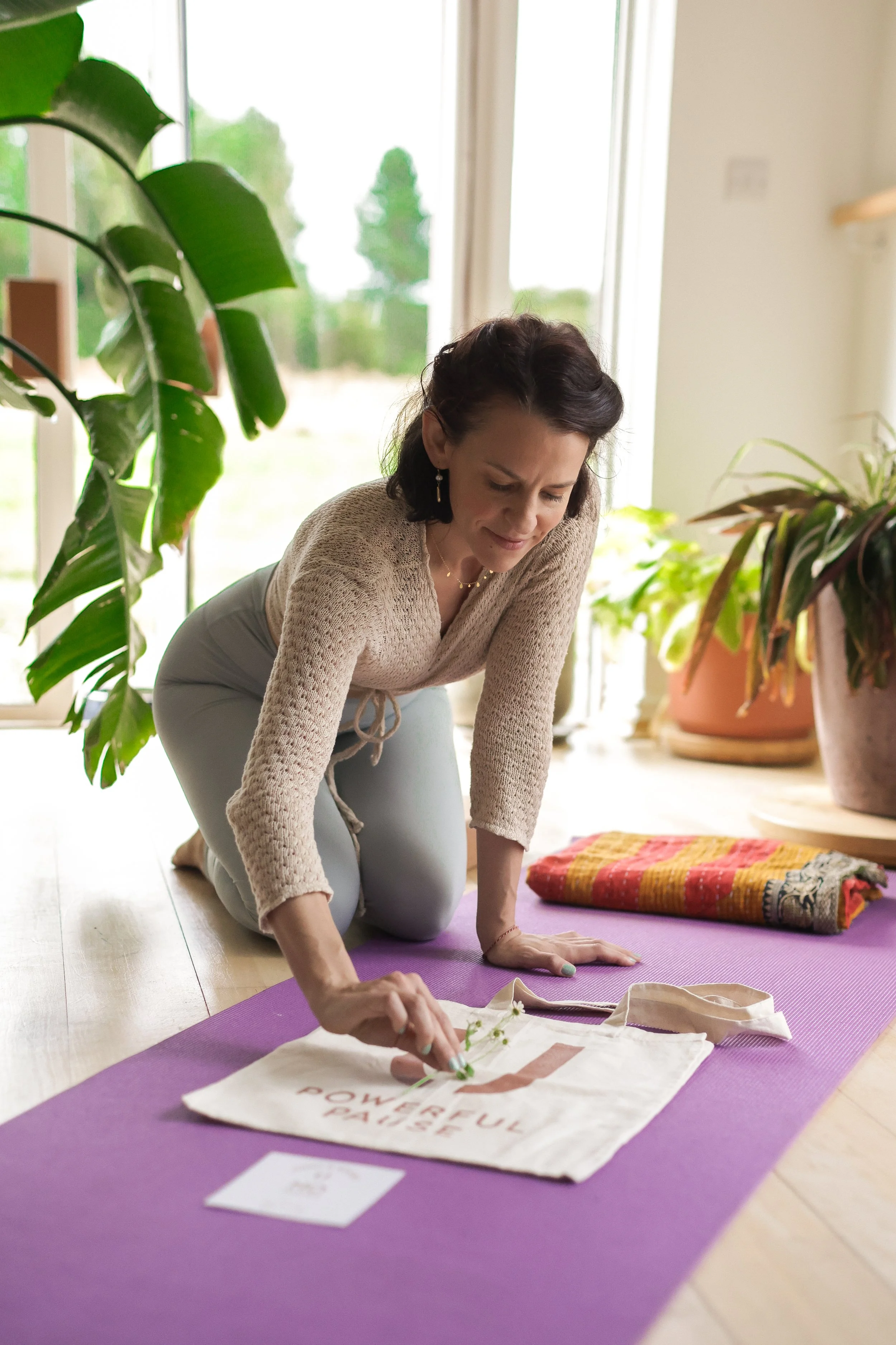 A woman practicing yoga on a purple mat in a sunlit room, surrounded by potted plants and a large window.