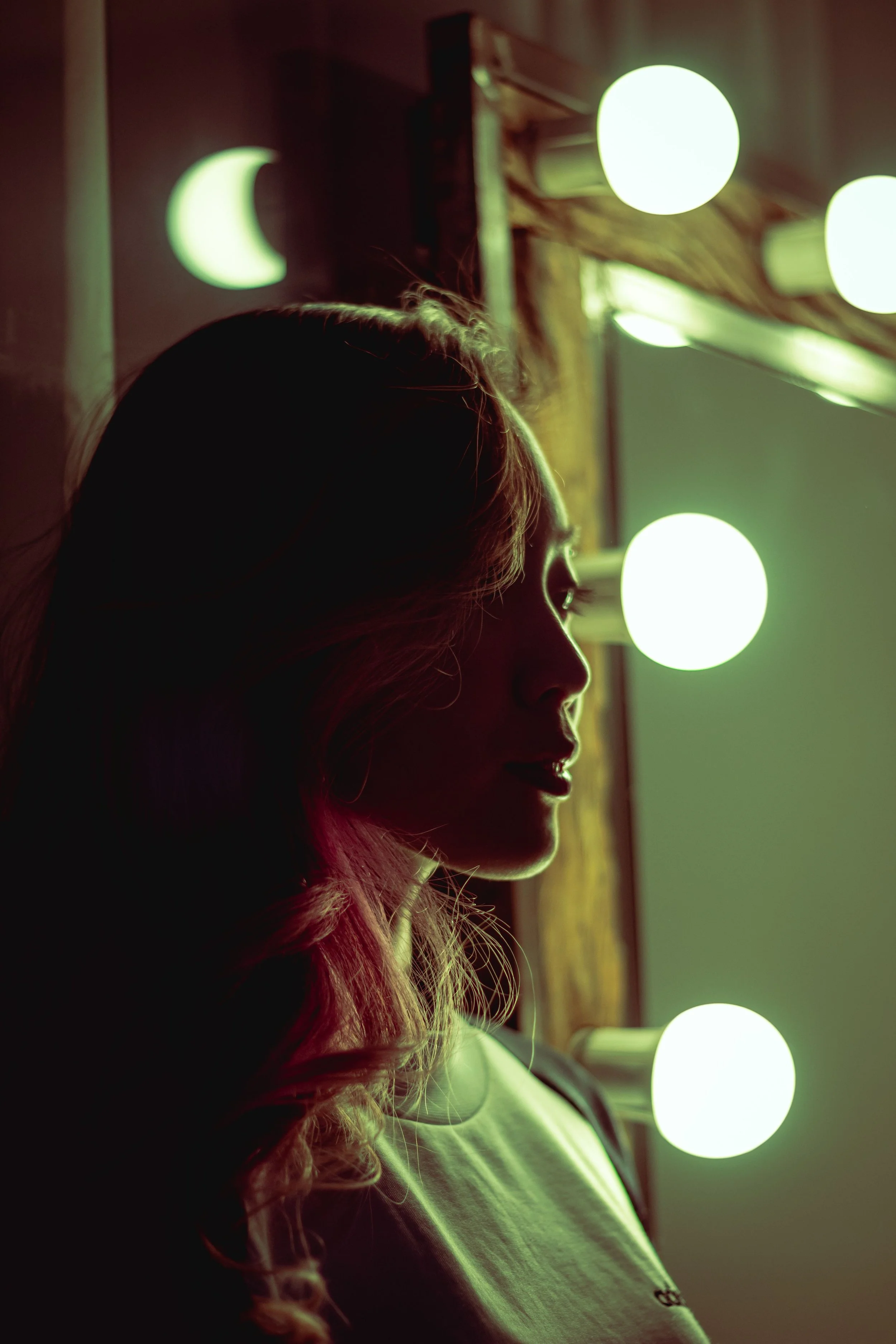 Silhouette of a woman with long hair looking at illuminated mirrors with round bulbs, in a room with warm lighting.