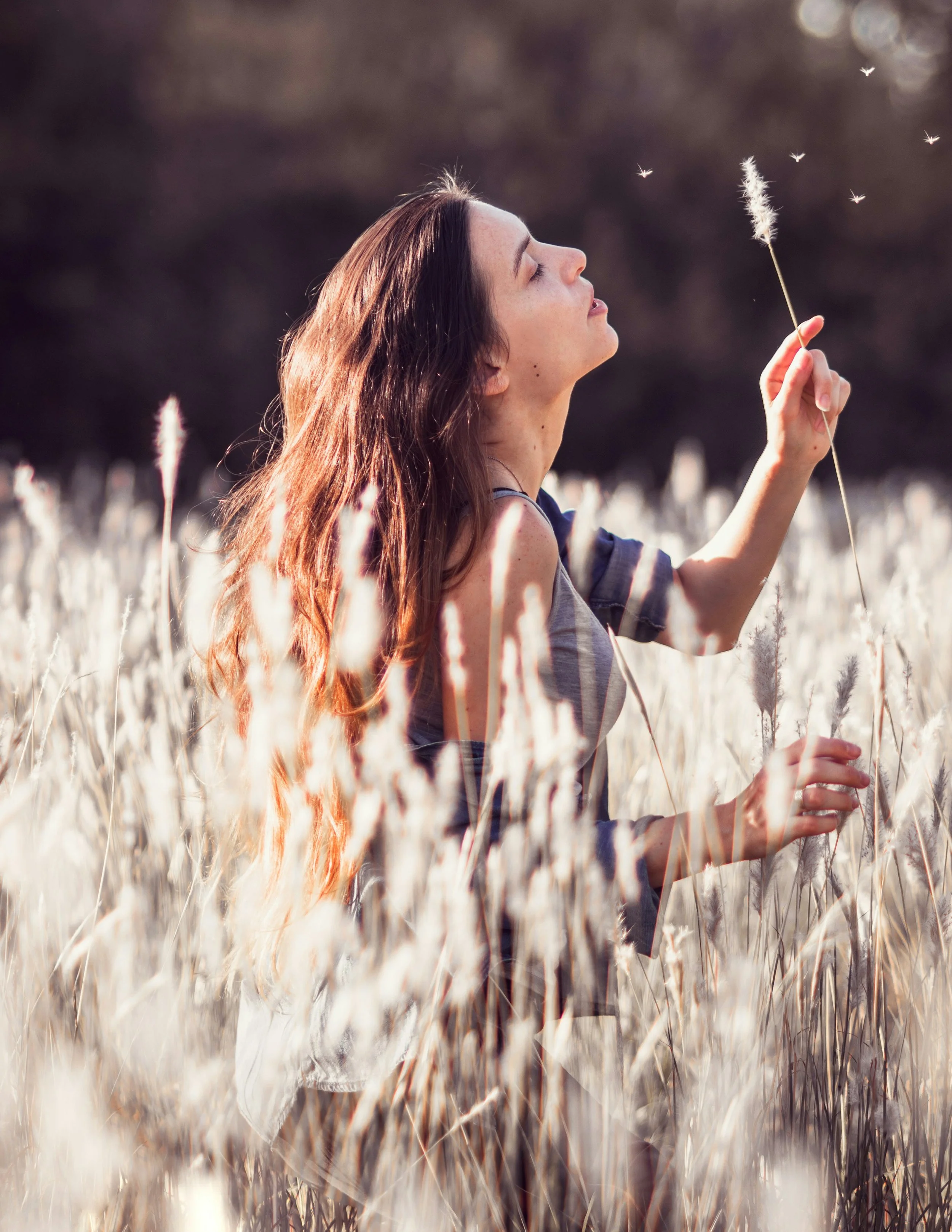 A woman with long brown hair in a field of tall white grass holding a single grass stalk and looking up with her eyes closed.