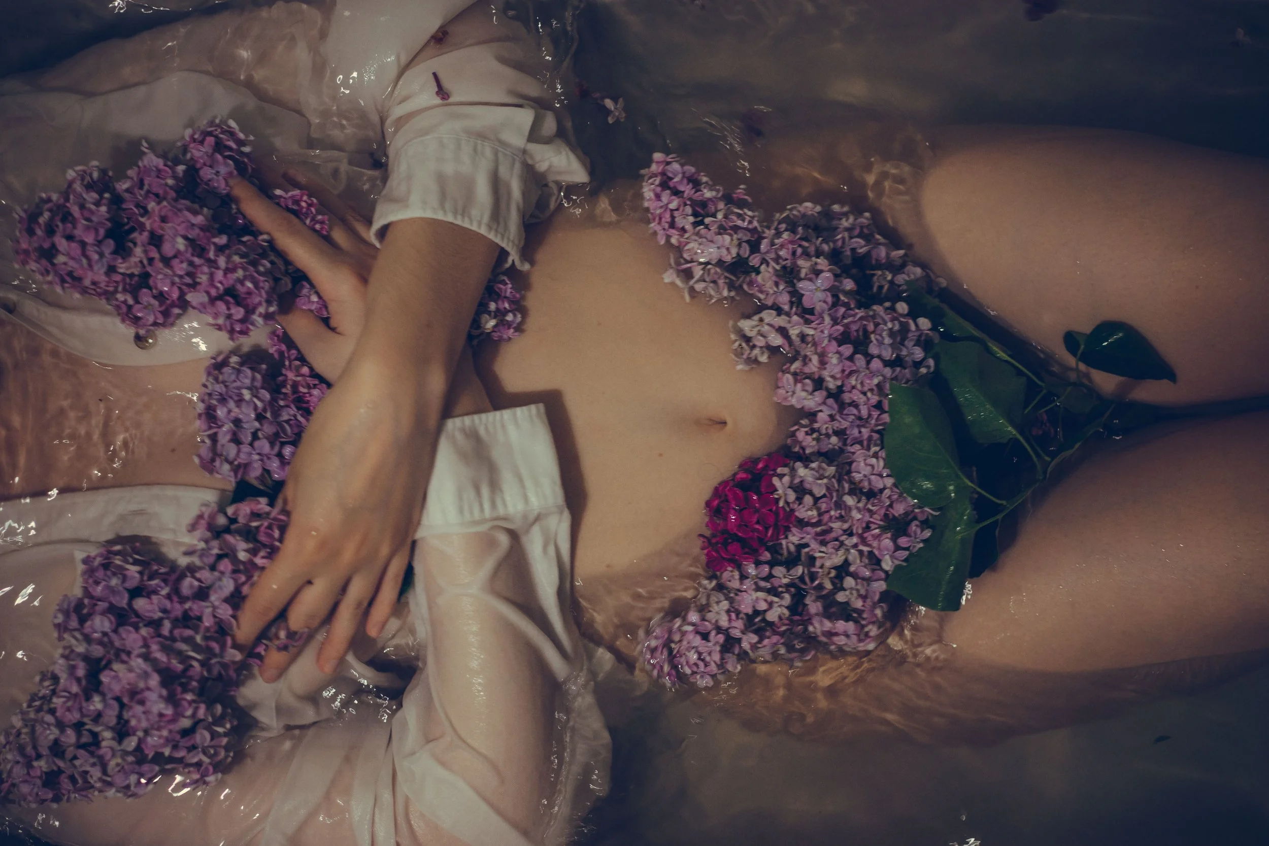 A naked woman lies in shallow water with flowers on her body