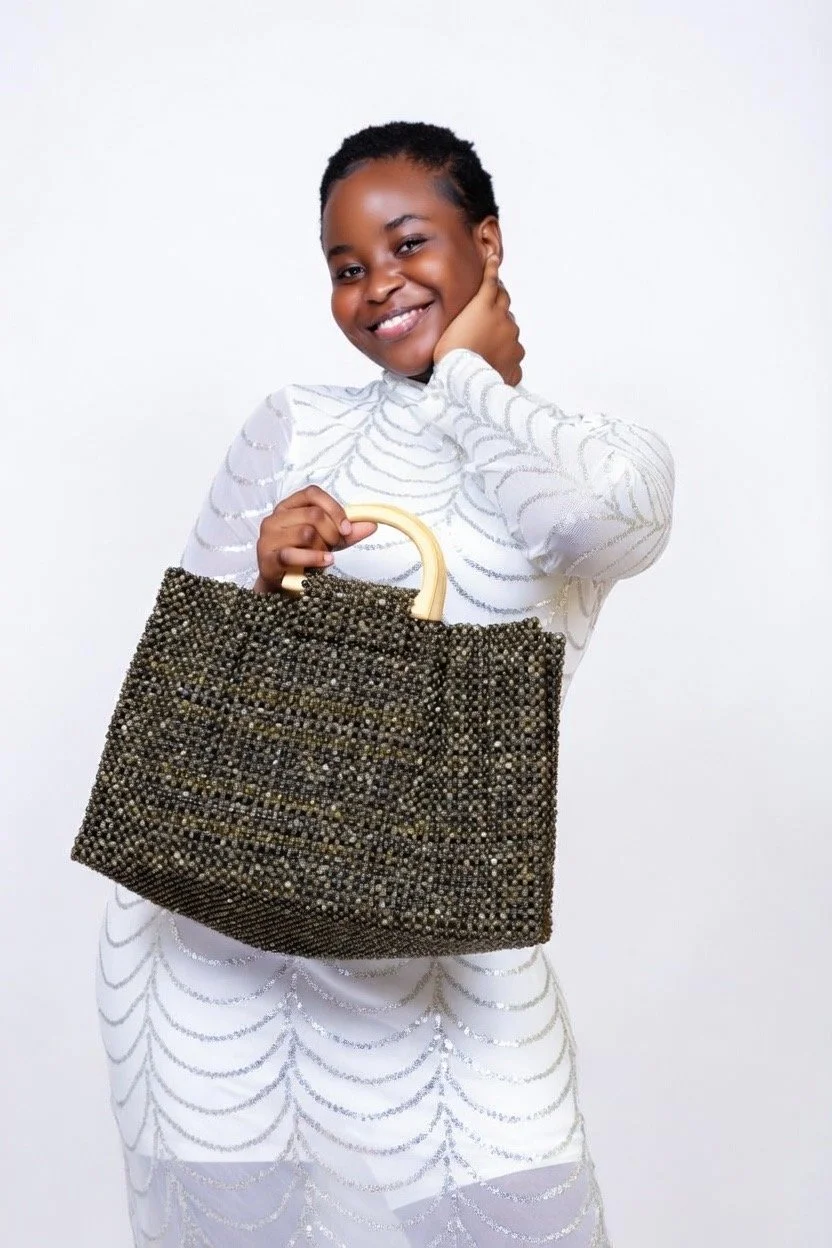 A woman smiling, holding a large, beaded handbag with wooden handles, wearing a black dress with silver sequin patterns, standing against a red background.