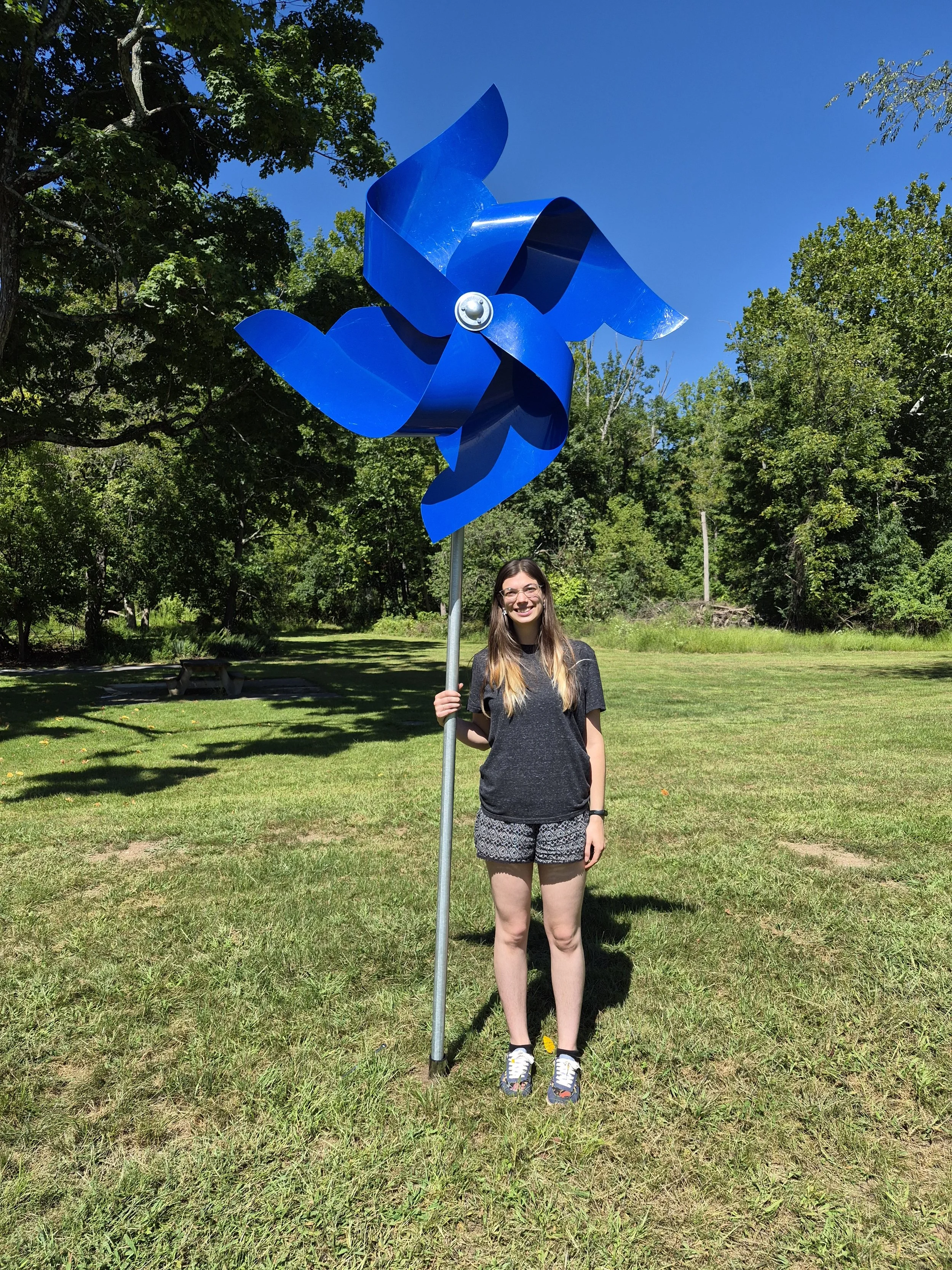 A giant blue pinwheel sculpture being held by the artists, a ombre haired woman with glasses, Maya Sorhage.